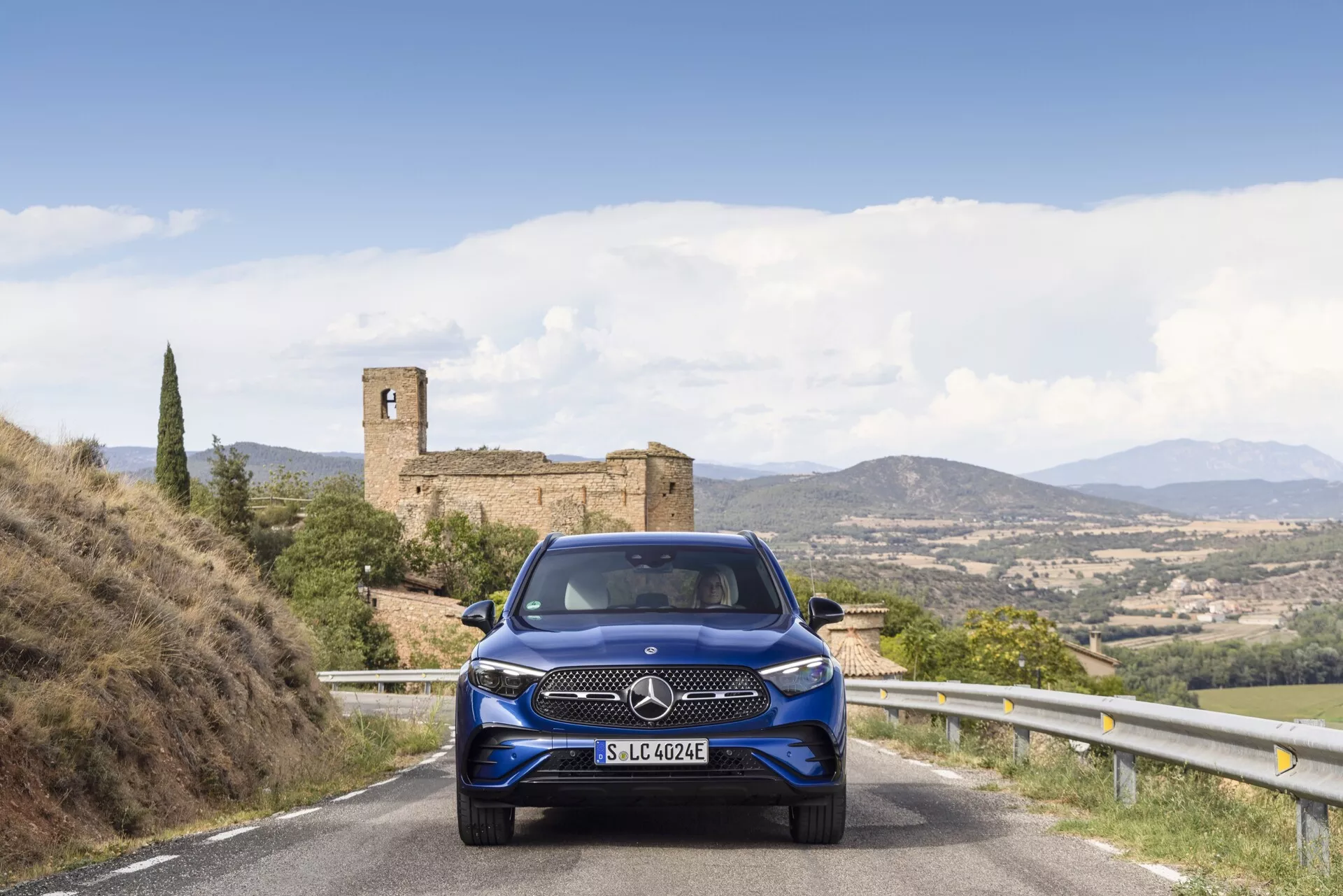 Mercedes-Benz GLC 254 de 2022 vu sous un angle avec la face avant et le côté visibles. La voiture dispose de phares LED élégants, une calandre proéminente et des jantes en alliage sportives. Garée dans une rue de la ville avec des bâtiments modernes en arrière-plan, sous une lumière du jour vive, mettant en évidence sa finition métallique argentée.