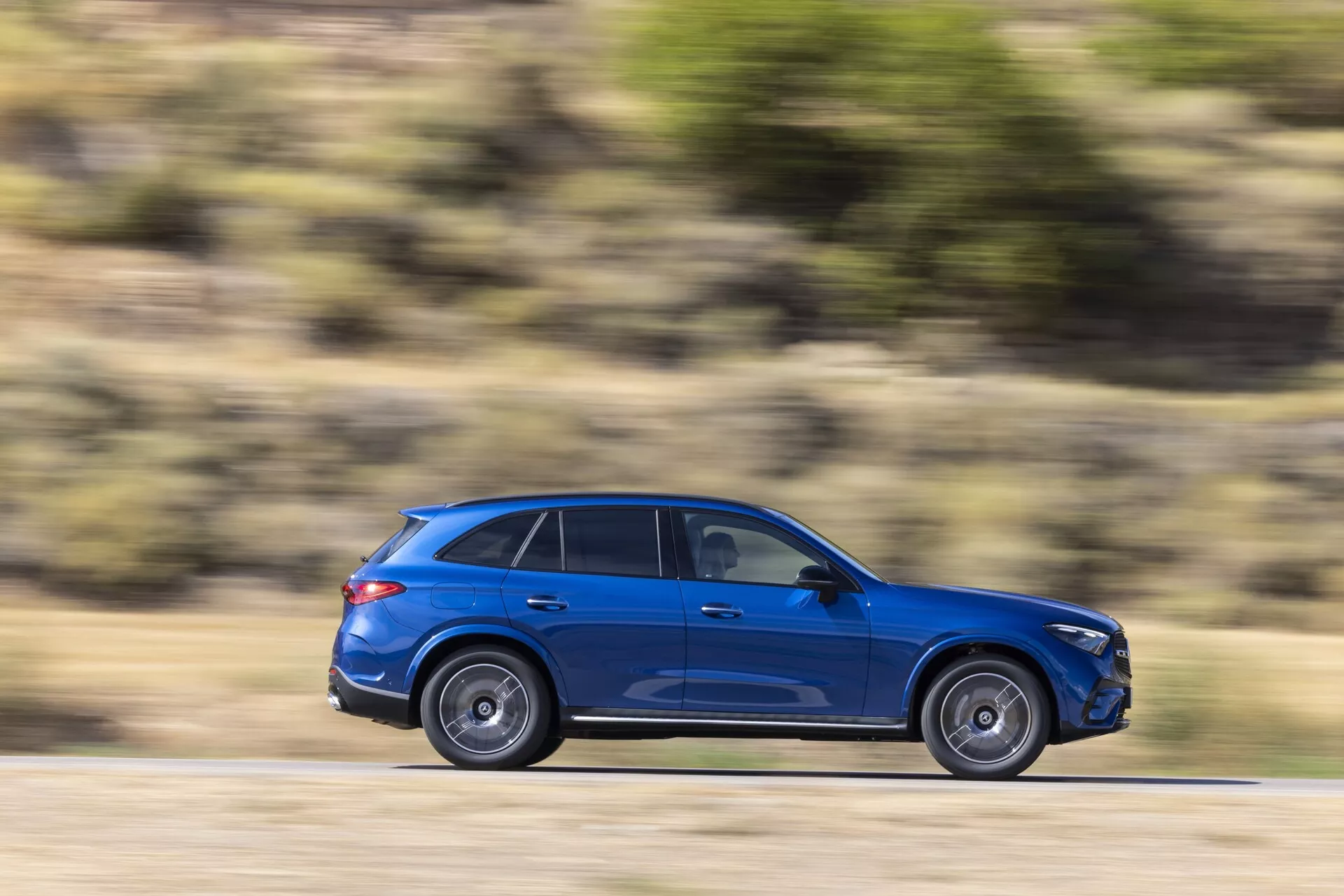 Profil latéral d'un SUV bleu vif en mouvement, capturé avec une technique de flou de mouvement accentuant la vitesse contre un arrière-plan strié horizontalement. Photo de jour avec éclairage naturel. Angle centré montrant toute la longueur du véhicule, design moderne et élégant, vitres teintées, surfaces réfléchissantes, silhouette de barres de toit et jantes floues indiquant le mouvement.