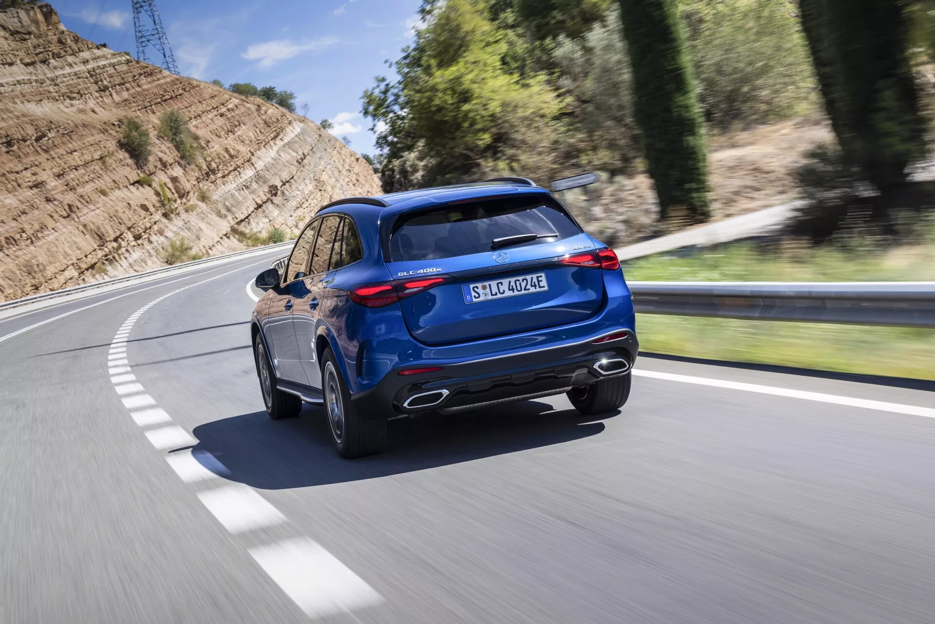 L'image présente une vue dynamique trois quarts arrière d'un SUV Mercedes GLC 2022 bleu roulant sur une route asphaltée tournant à gauche. Capturé sous un angle légèrement surélevé, les points forts du véhicule incluent des embouts d'échappement chromés, un pare-chocs arrière substantiel avec des garnitures noires, des feux arrière à LED élégants et une lunette arrière fortement inclinée. Le spoiler de toit sportif, le toit ouvrant panoramique et les barres de toit visibles ajoutent à l'attrait haut de gamme. Le profil latéral révèle des jantes en alliage multibrins et des pneus à profil bas, mettant l'accent sur les performances routières. Une prise de charge devant le feu arrière et un badge EQ indiquent des capacités hybrides rechargeables ou électriques, tandis que le badge 4MATIC indique une traction intégrale. Le flou de mouvement en arrière-plan suggère que le véhicule est en mouvement, offrant une impression de vitesse et de mouvement.
