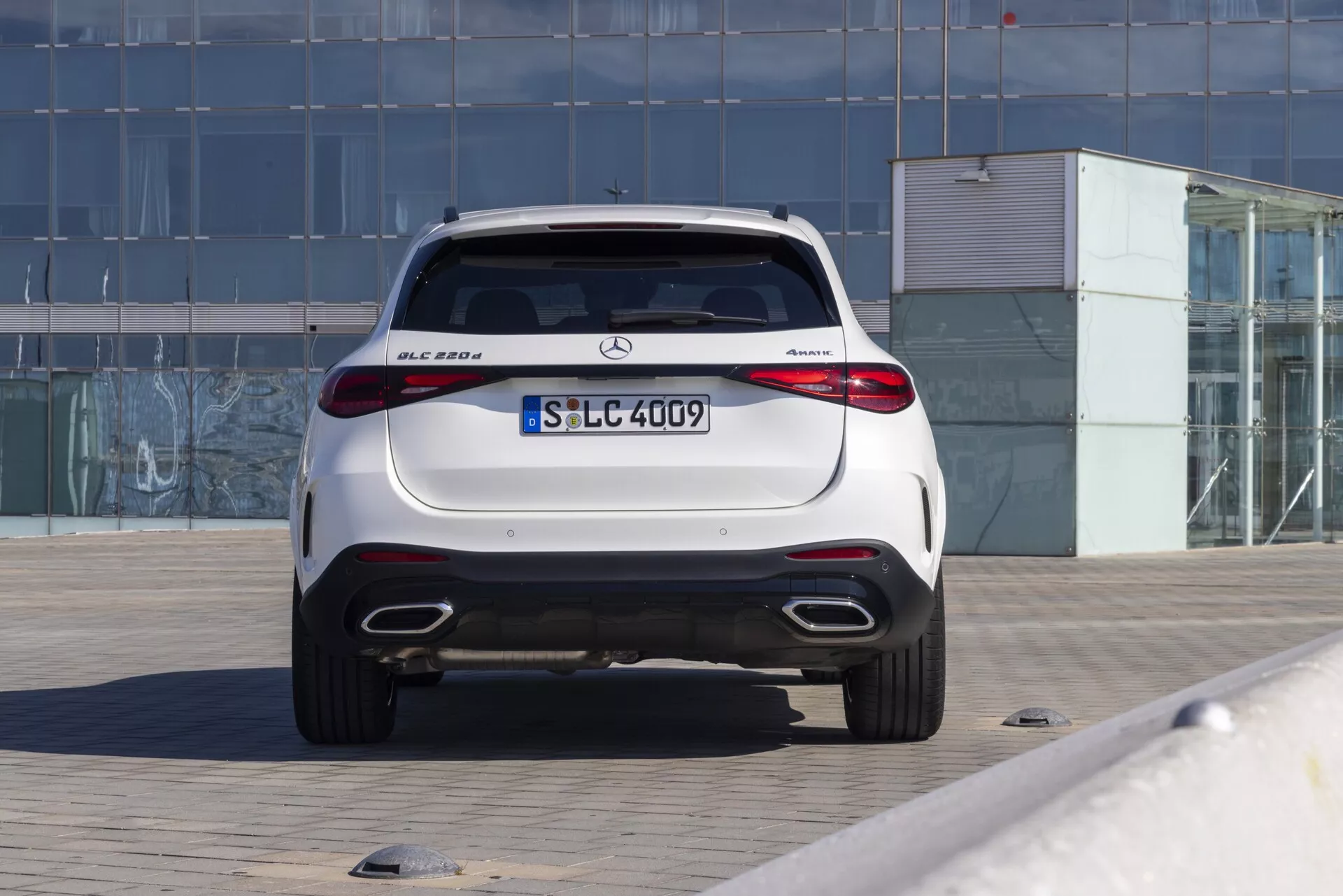 Vue arrière d'un SUV compact de luxe Mercedes-Benz GLC 300 de 2022 avec capacité de traction intégrale 4MATIC, garé sur une zone pavée. La photographie montre le hayon aérodynamique, le spoiler intégré, les feux arrière LED au design horizontal, les doubles sorties d'échappement, les capteurs de stationnement intégrés, le diffuseur arrière et la finition brillante. La voiture est centrée dans le cadre avec un fond montrant un bâtiment avec une façade en verre, indiquant un environnement urbain ou commercial. L'image bien éclairée met en évidence les caractéristiques de design élégant et sportif typiques des véhicules modernes Mercedes-Benz.