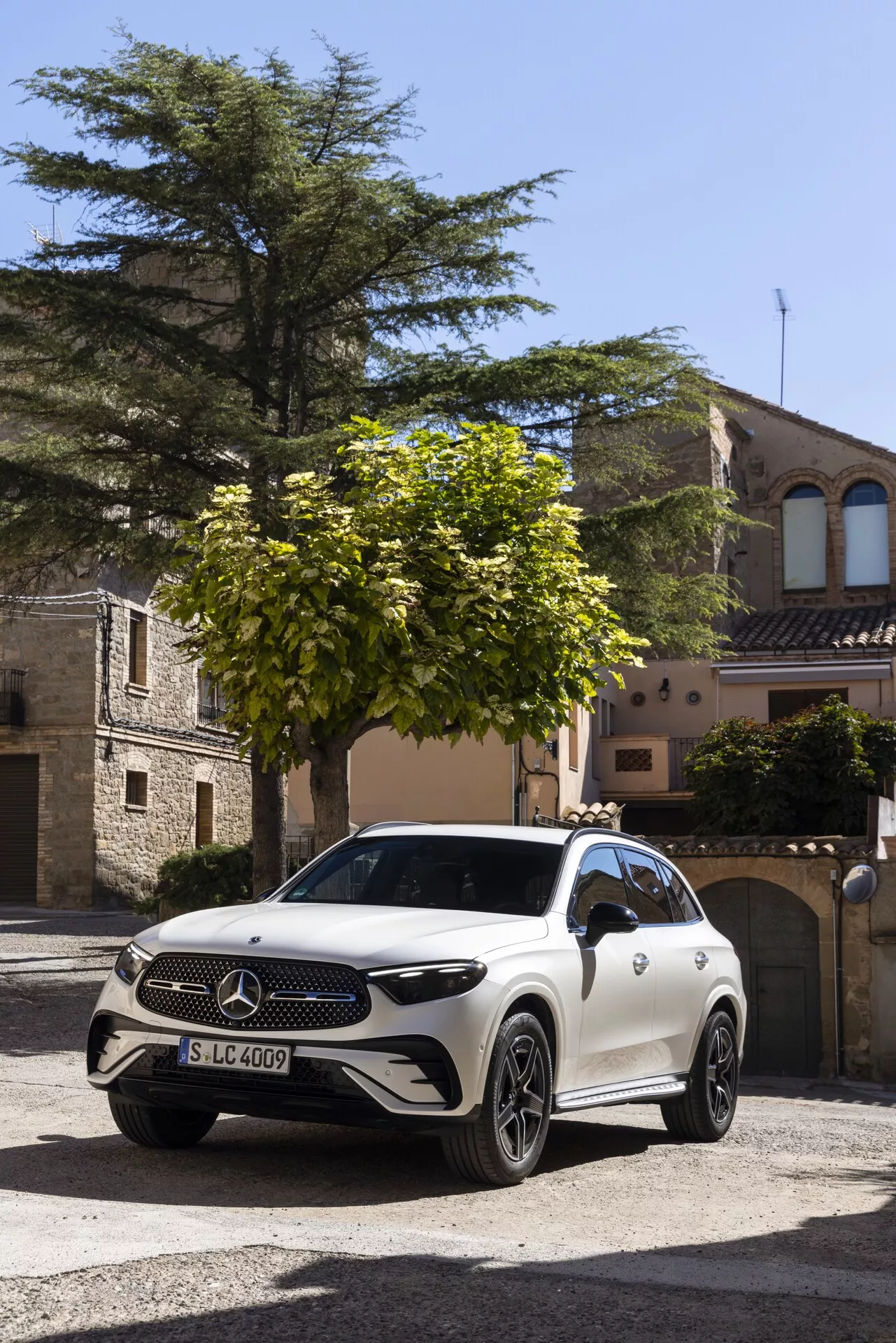 SUV Mercedes-Benz GLC 2022 (254) blanc stationné sur une rue pavée dans une ville européenne historique, pris sous un angle frontal trois-quarts. Les caractéristiques visibles incluent un grand emblème Mercedes-Benz sur la calandre, des phares LED, des jantes en alliage, des caméras/senseurs potentiels sur le pare-chocs avant, un design aérodynamique et des rétroviseurs extérieurs pouvant loger des caméras supplémentaires ou des indicateurs de direction. L'environnement et la lumière naturelle soulignent l'apparence luxueuse du véhicule.