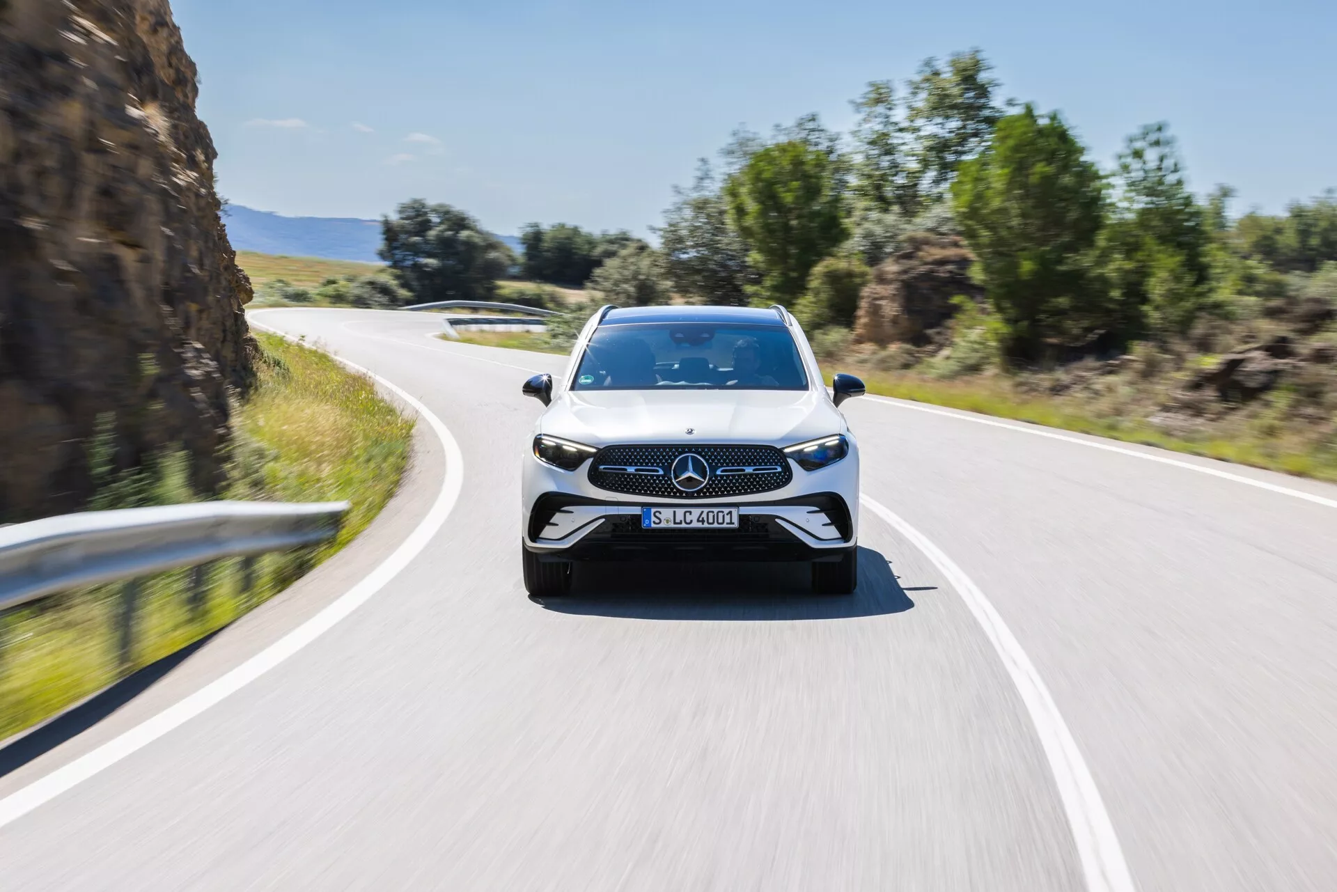 Vue de face d'une Mercedes-Benz GLC 2022 en mouvement, capturée légèrement décentrée, montrant l'avant et une partie des côtés. L'image met en avant l'emblème et le design élégant de la calandre, les phares LED, les grandes prises d'air et les lignes sculptées du capot, suggérant un design sportif et moderne. L'arrière-plan flou indique une sensation de vitesse, avec la voiture circulant sur une route à deux voies sous un ciel dégagé, accentuant ses caractéristiques dynamiques et technologiques avancées.