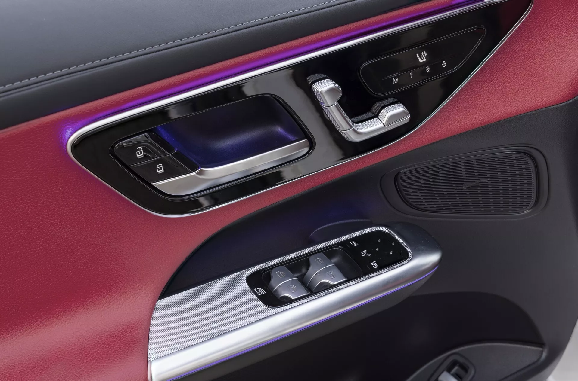 Gros plan du panneau de porte côté conducteur de la Mercedes GLC (254) 2022, présentant un mélange de matériaux, notamment des garnitures métalliques et des finitions noires mates. Comprend un éclairage d'ambiance coloré, une poignée de porte ergonomique en noir brillant et métal, des commandes électriques des fenêtres rétroéclairées, des rétroviseurs extérieurs réglables électriquement et une grille de haut-parleur haute fidélité intégrée en bas, mettant en valeur le design intérieur luxueux et technologiquement avancé de la voiture.