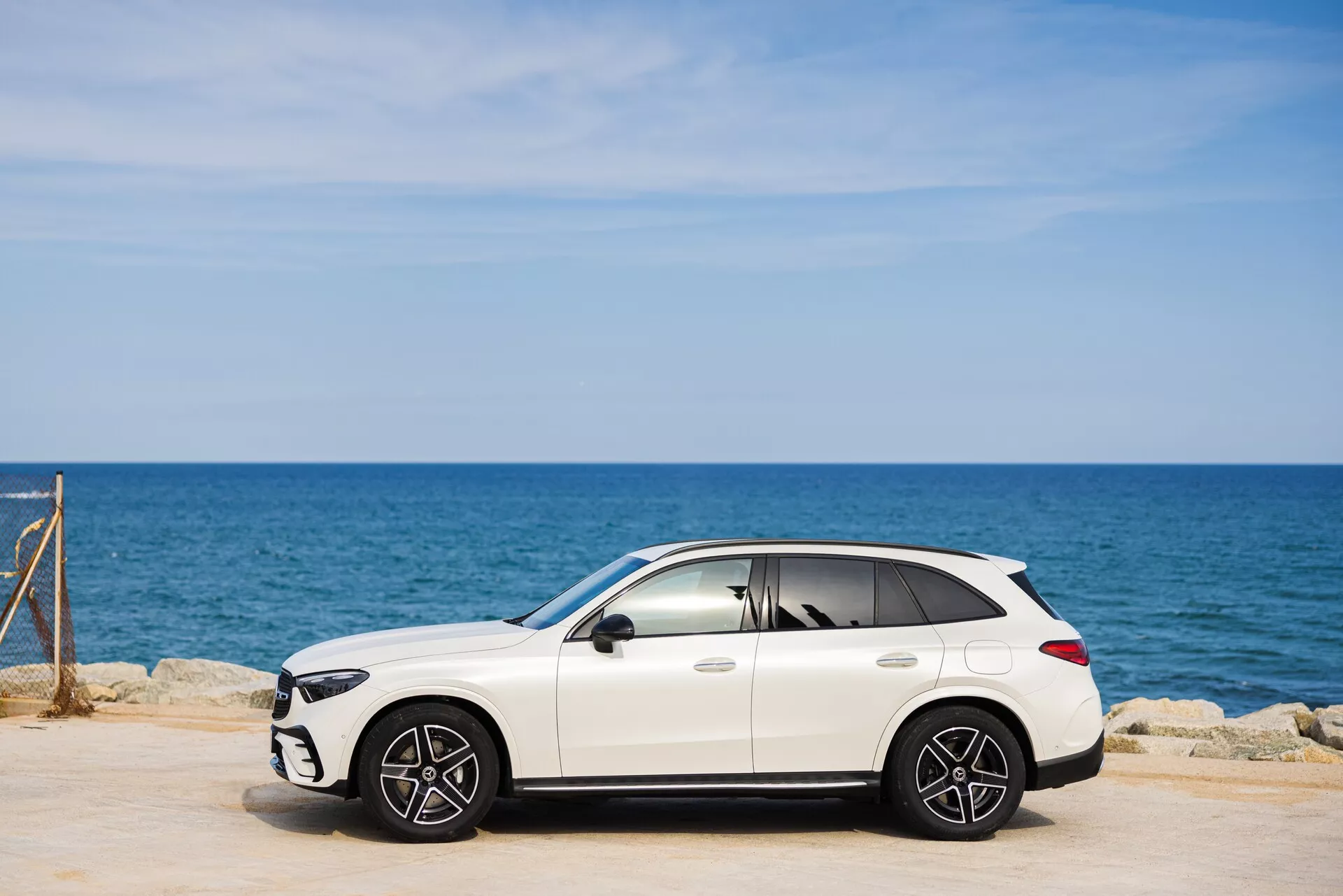 Image d'un SUV crossover de luxe Mercedes GLC 2022 (254) blanc stationné sur une zone pavée. La perspective de côté met en valeur le profil du côté passager avec les roues avant et arrière à plusieurs rayons visibles. En arrière-plan, la mer et un ciel bleu clair. Le design élégant du SUV comprend des rétroviseurs, des poignées de porte et un éventuel toit ouvrant. Les ombres sous le véhicule indiquent des conditions ensoleillées. Les fenêtres légèrement teintées ajoutent un aspect luxueux.