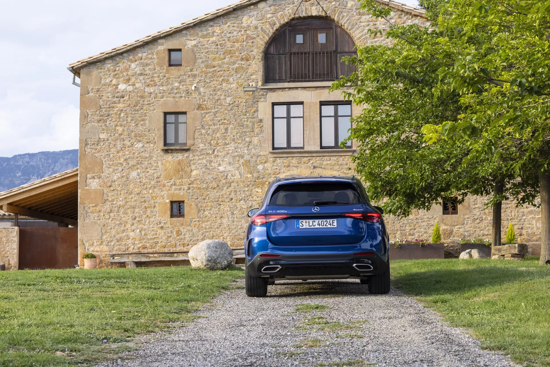 Vue arrière d'une Mercedes GLC 2022 bleue (254) stationnée sur une allée de gravier, centrée dans la composition avec un bâtiment en arrière-plan. L'image met en évidence le hayon, le pare-brise arrière avec essuie-glace, les feux arrière LED élégants, le pare-chocs arrière avec capteurs de stationnement ou réfléchisseurs et le système d'échappement double. Les caractéristiques comprennent un feu stop en hauteur, une plaque d'immatriculation centralisée, un élément de diffuseur et un aileron de toit, montrant la technologie et le design automobile moderne.
