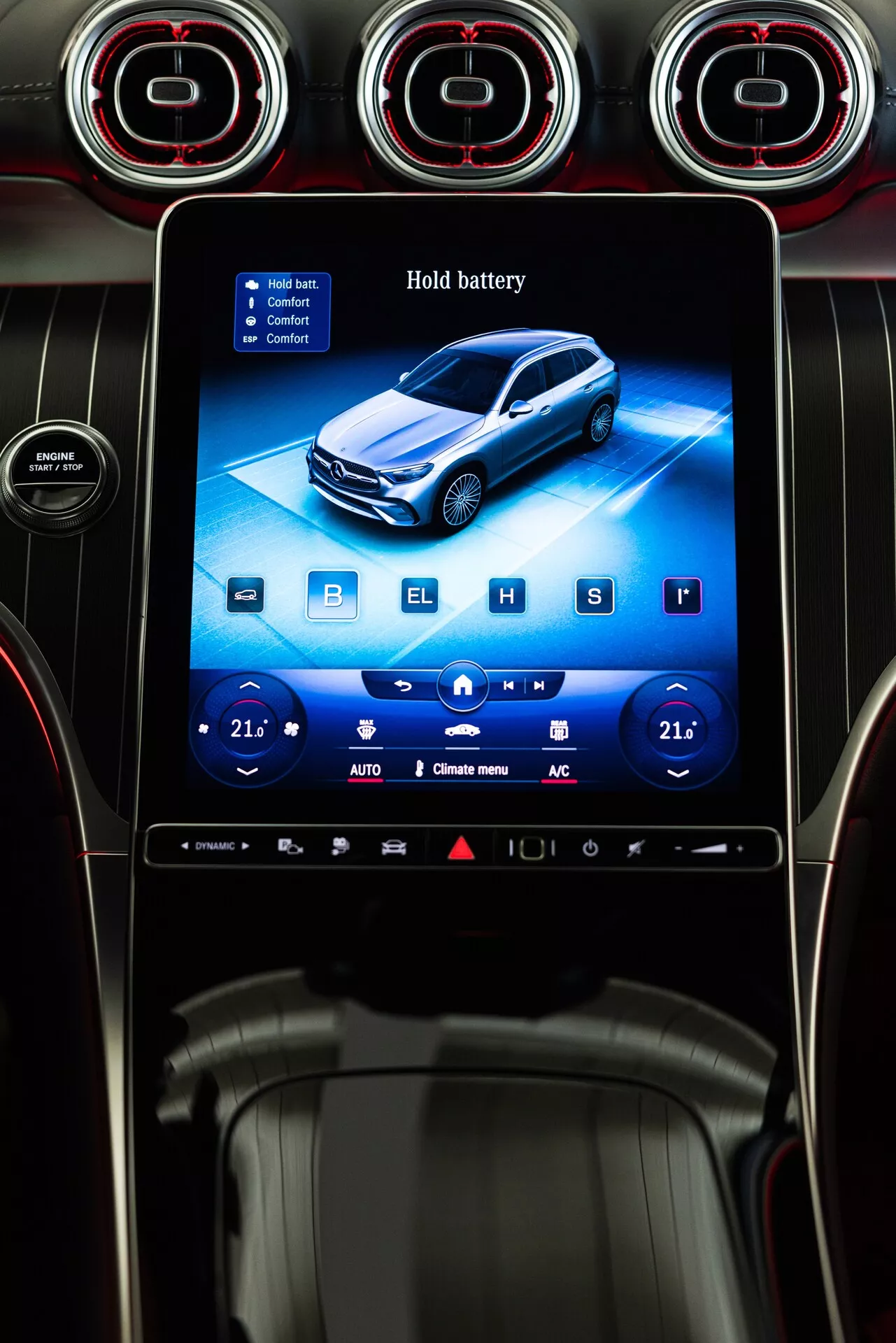 Photographie de l'intérieur d'une Mercedes GLC 254 de 2022, mettant en évidence un écran d'infodivertissement rectangulaire verticalement orienté, situé au centre du tableau de bord. L'écran, entouré d'un entourage noir brillant, présente une interface thématique bleue, possiblement indiquant des fonctionnalités hybrides ou électriques, avec la fonction 'Hold battery' visible. L'interface inclut des commandes tactiles pour la climatisation, la lecture multimédia et les paramètres de la voiture, avec des boutons étiquetés 'B', 'E', 'H' et 'S' pour différents modes de conduite ou paramètres de la batterie. Au-dessus de l'écran, trois bouches d'aération circulaires avec des accents rouges indiquent un design premium. La partie inférieure de l'écran présente des commandes de climatisation double zone réglées à 21°C, avec des icônes pour le mode auto, la circulation de l'air et la climatisation, ajoutant à l'esthétique moderne et haut de gamme. La scène révèle également une bordure argentée étroite et une partie de la zone du levier de vitesse, avec un aperçu d'un porte-gobelet ou compartiment de rangement.
