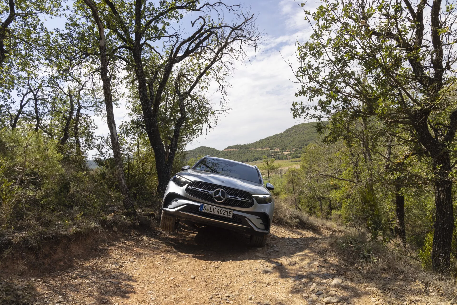 Image en couleur d'un SUV Mercedes-Benz GLC 2022 dans un cadre naturel, capturée sous un angle frontal bas, mettant en valeur son aspect robuste. La photo se concentre sur la partie avant de la voiture, la grille et les phares LED modernes. Le véhicule apparaît sur un chemin de terre, entouré de feuillage, mettant en évidence ses capacités tout-terrain et son design élégant. Les capteurs et caméras avant suggèrent des fonctionnalités de sécurité avancées.