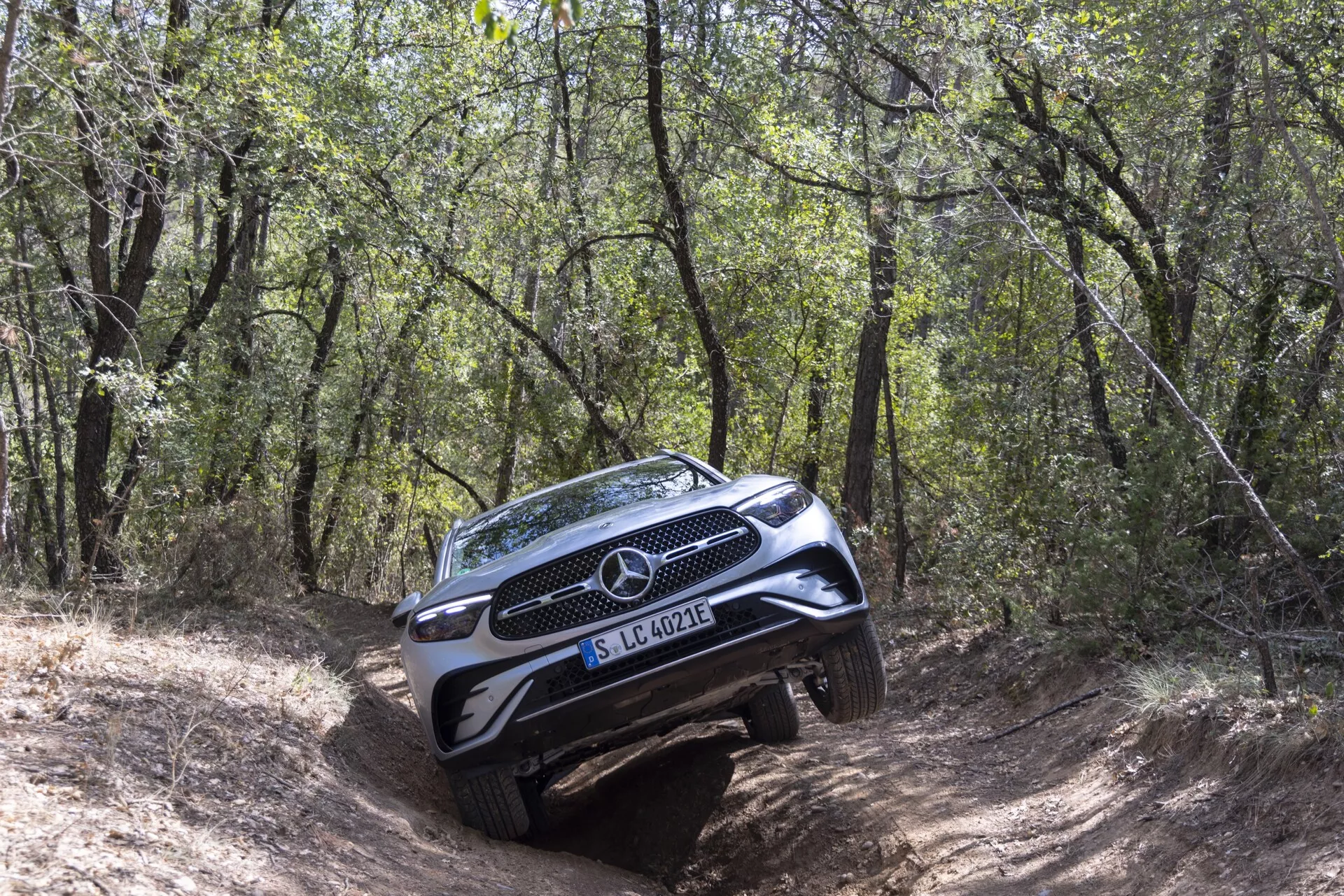 SUV Mercedes-Benz GLC 2022 naviguant sur un sentier hors route boisé et difficile, vu d'un angle frontal de trois quarts, montrant la calandre complète, l'emblème, les phares, une partie du capot et le pare-chocs avant avec plaque de protection; côté passager plus visible avec une position inclinée sur un terrain inégal, entouré d'arbres et de buissons, capturant les capacités hors route du SUV.