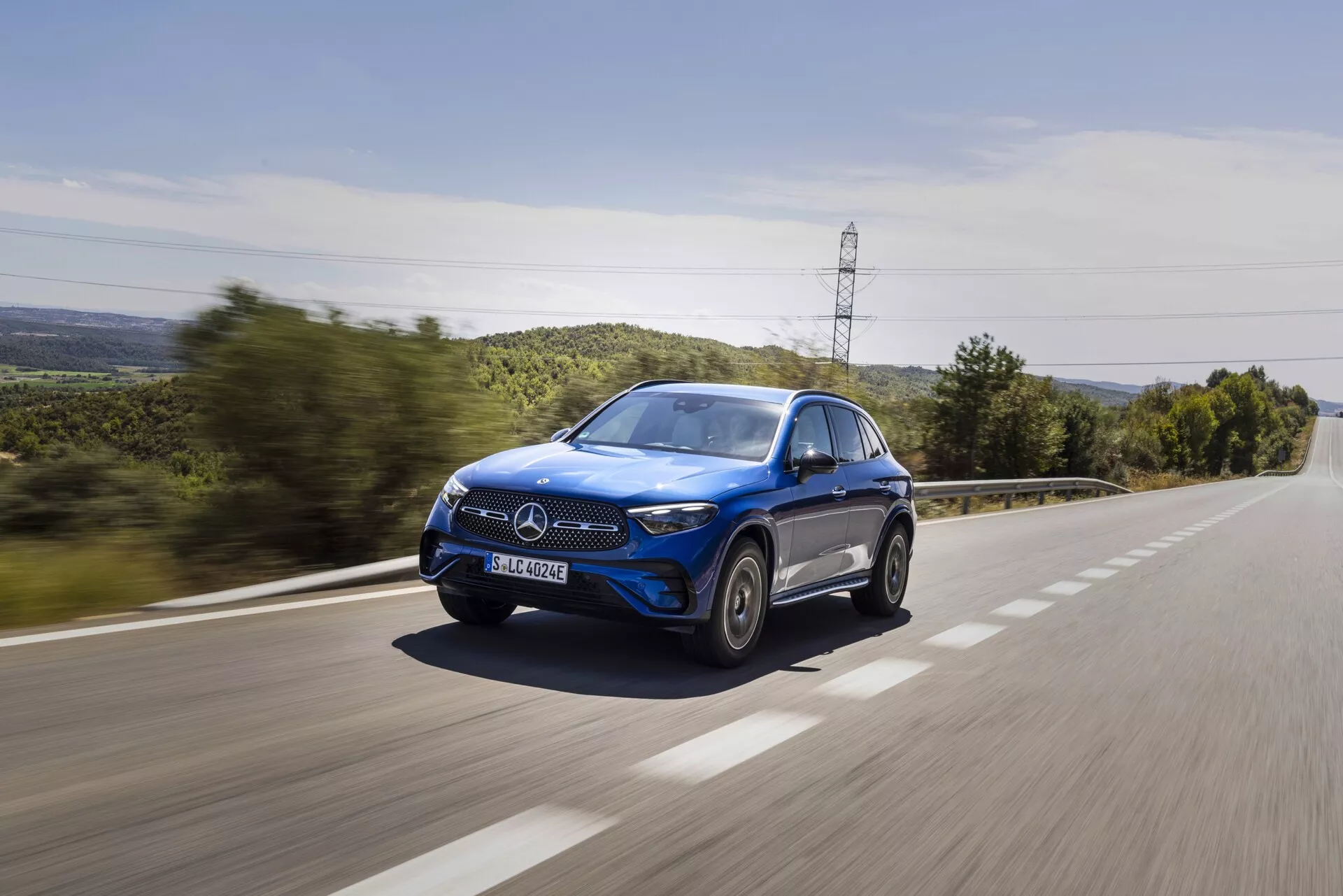 Un SUV bleu Mercedes-Benz GLC 2022 roulant à grande vitesse sur une route de campagne, capturé sous un angle de trois quarts avant, montrant la calandre avant, le capot, les phares LED, le rétroviseur côté conducteur, les roues et les fenêtres, avec un effet de flou de mouvement sur la surface de la route et la verdure en arrière-plan, mettant en valeur le design dynamique et luxueux de la voiture.