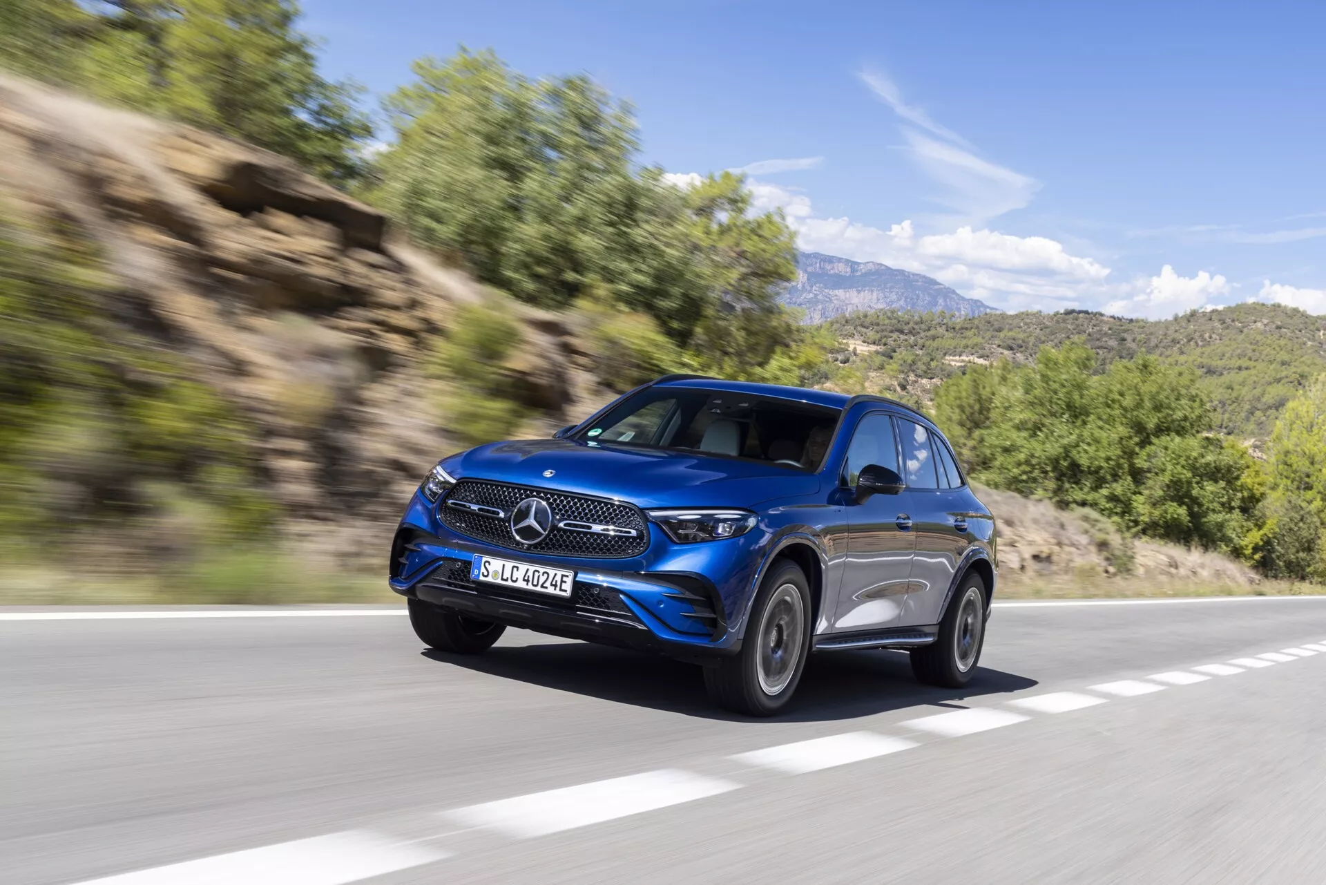 Prise dynamique d'un SUV bleu Mercedes GLC 2022 (254) en mouvement, vu de l'angle avant gauche. L'image montre ses phares élégants, sa calandre proéminente avec emblème central, ses roues en alliage haut de gamme, la ligne aérodynamique de la carrosserie et ses rétroviseurs latéraux avancés. Le véhicule, roulant sur une route goudronnée dans une zone rurale montagneuse, se détache sur un fond de feuillage et de collines rocheuses floues par le mouvement, ajoutant une impression de vitesse et de luxe moderne.