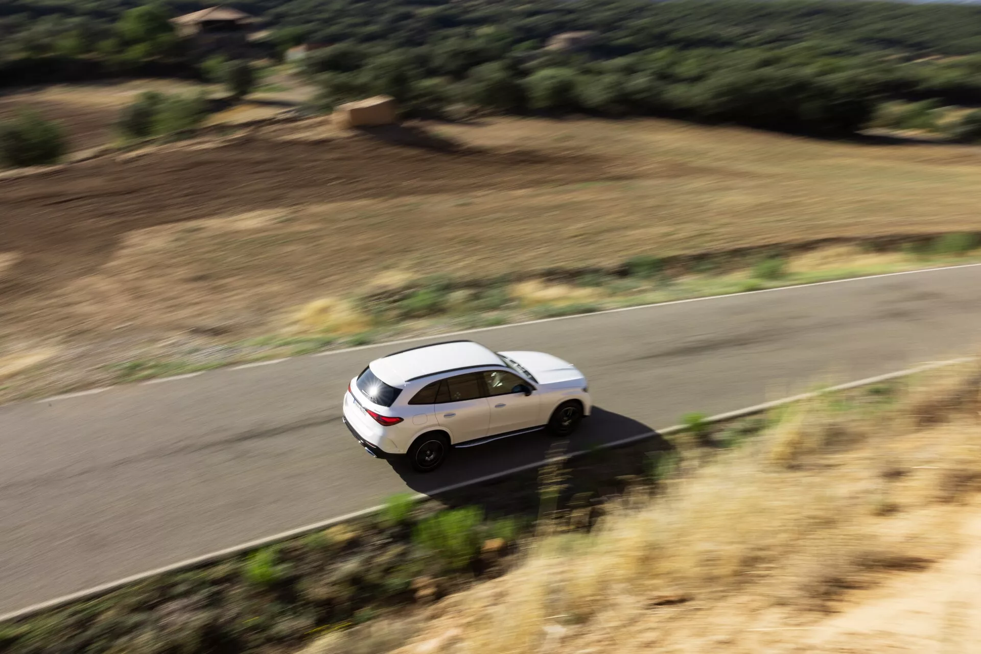 Vue en hauteur d'un SUV Mercedes GLC 2022 blanc à quatre portes en mouvement sur une route sinueuse en milieu rural, entourée par un paysage naturel. Le véhicule présente une carrosserie aérodynamique moderne, un toit ouvrant panoramique, des jantes en alliage, des vitres teintées à l'arrière et des rétroviseurs latéraux avec clignotants intégrés. Le flou de mouvement suggère la vitesse et une performance dynamique.