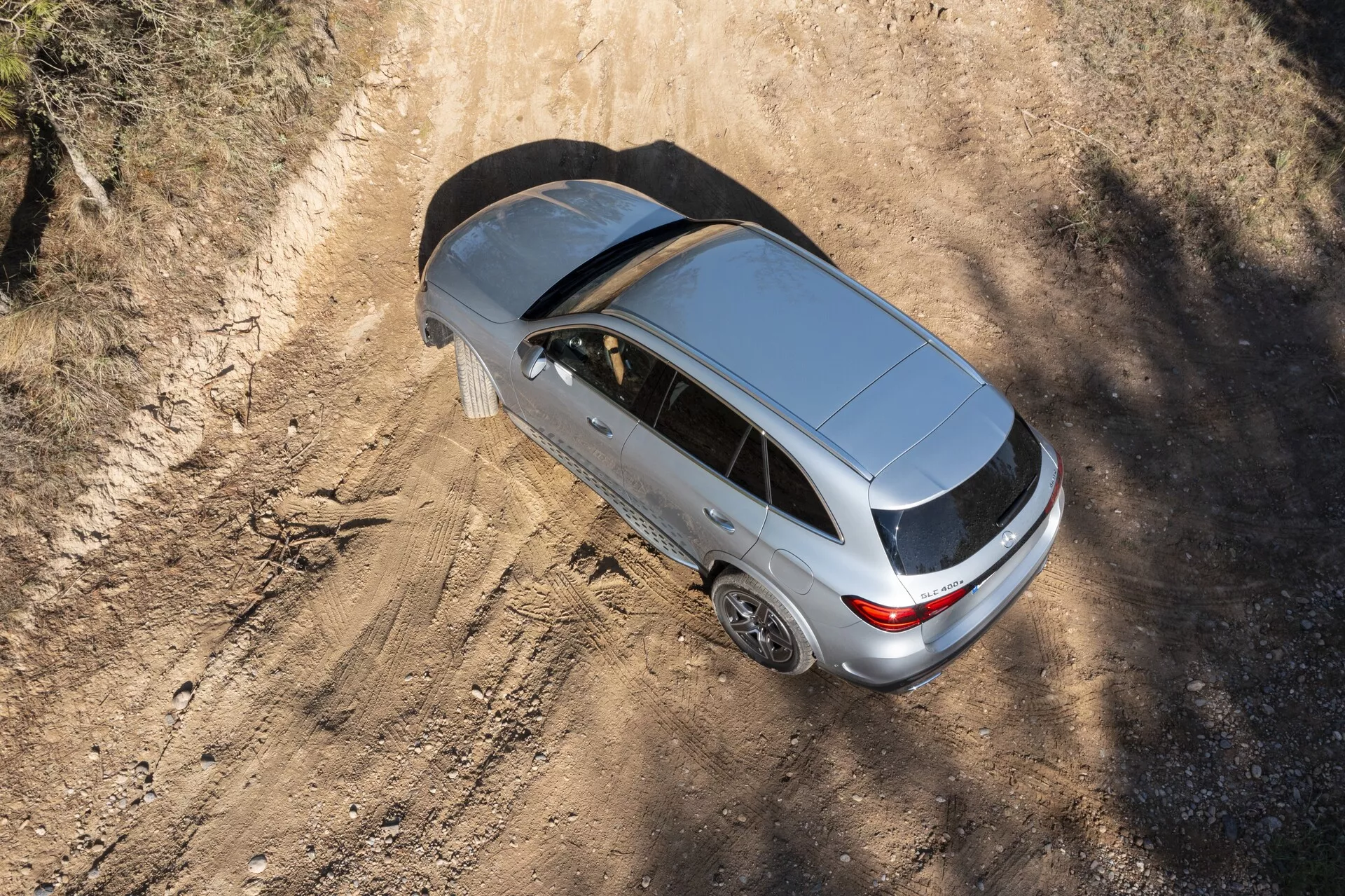 Vue aérienne d'une Mercedes GLC 2022 argentée, présentant son design moderne avec un toit ouvrant fermé, une antenne arrière et des jantes en alliage à cinq branches. La voiture, positionnée sur un chemin rural poussiéreux, met en avant son possible style de carrosserie hatchback ou sportback, avec des ombres indiquant la lumière du matin ou de l'après-midi.