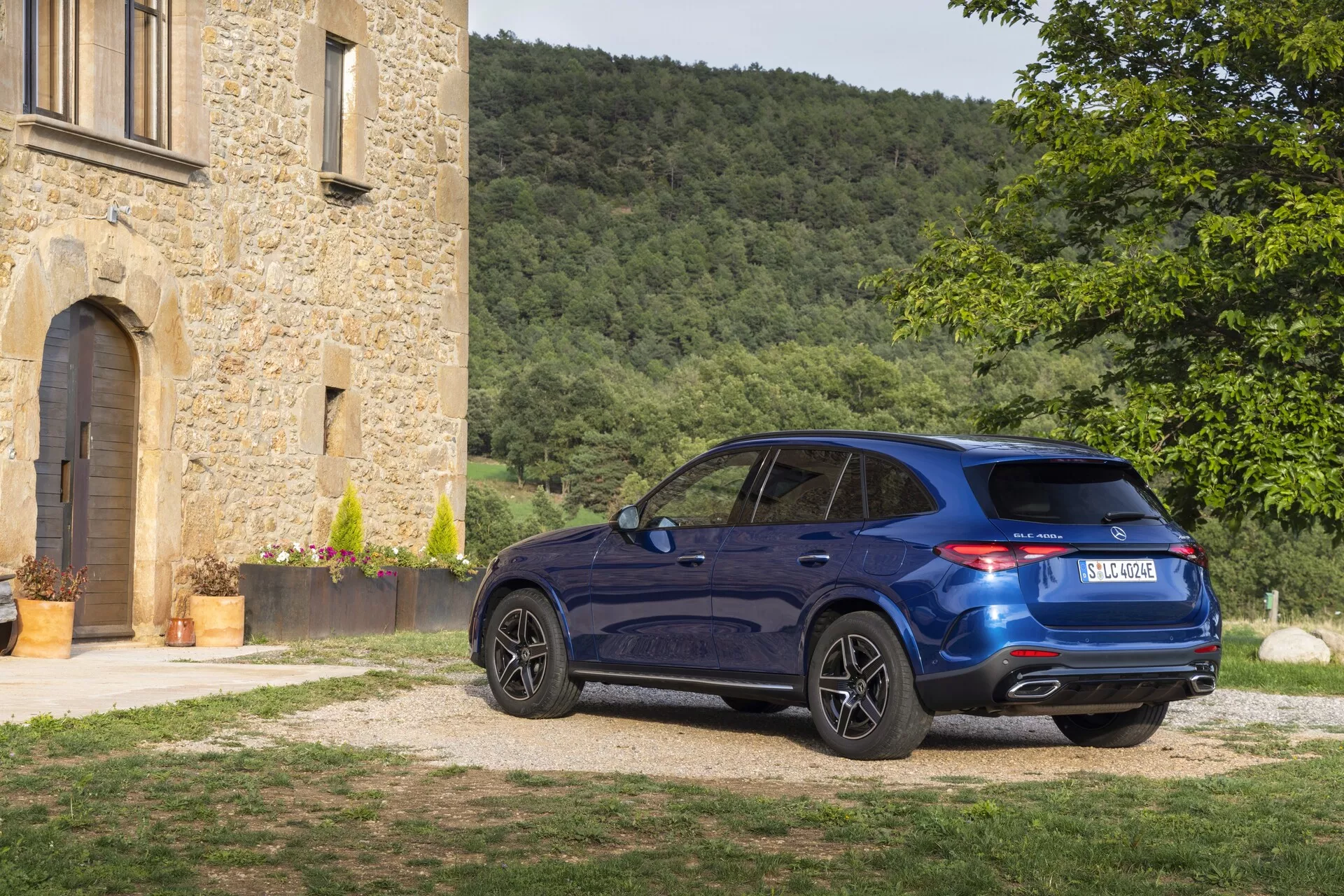 SUV crossover de luxe Mercedes-Benz GLC 2022 sur une allée en gravier près d'un bâtiment en pierre historique, vu sous une perspective arrière trois-quarts, montrant la peinture bleu profond, les feux arrière LED, le système d'échappement double, les jantes en alliage et la ligne de toit sportive sous la lumière de midi.