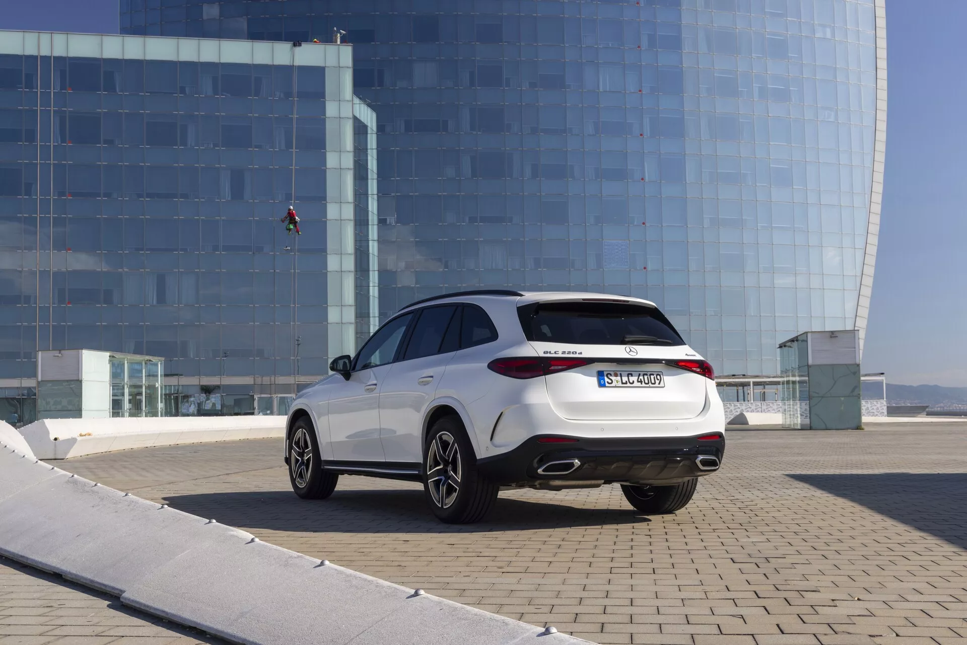 Sur l'image, une voiture moderne Mercedes GLC 2022 (254) est capturée avec un angle arrière trois-quarts extérieur, positionnée en diagonale avec l'arrière plus proche de la caméra, montrant des feux arrière LED contemporains, un système d'échappement double, des vitres arrière teintées, une ligne de toit descendant en douceur, des jantes en alliage multi-branches avec des pneus à profil bas, le tout dans un cadre urbain avec un bâtiment en verre réfléchissant en arrière-plan.