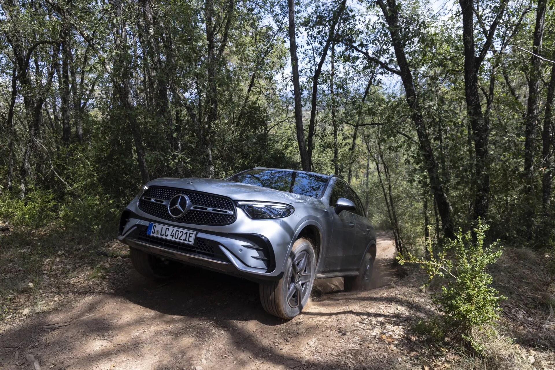 SUV Mercedes GLC 2022 positionné sur un chemin de terre entouré d'une zone boisée, capturé sous un angle trois-quarts avant mettant en valeur sa calandre avant proéminente avec l'emblème de la marque, les phares à LED, les roues en alliage et une hauteur de caisse élevée, mettant en avant ses capacités tout-terrain dans un environnement naturel d'arbres et de terrain sec.