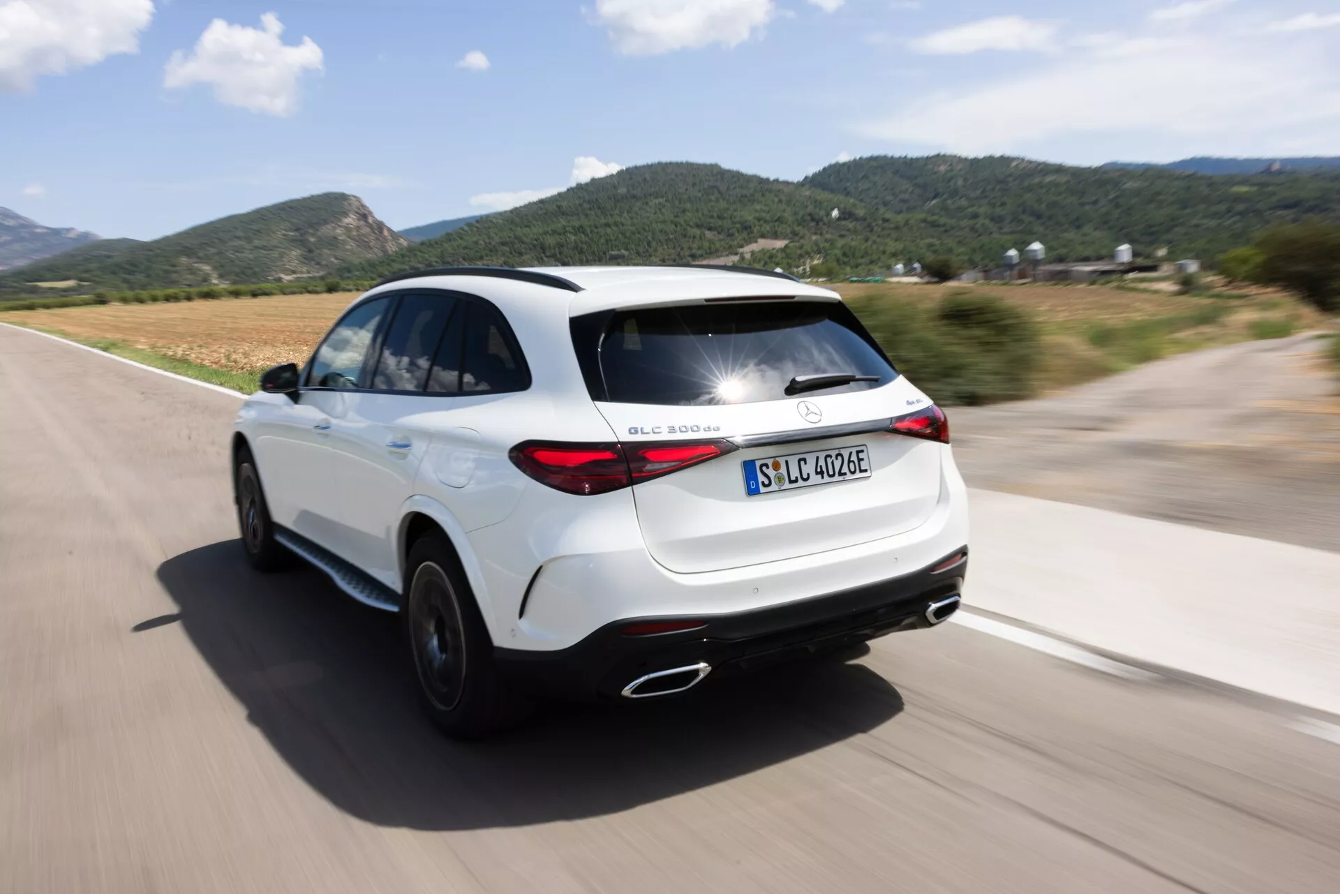 Vue arrière dynamique des trois quarts d'un SUV blanc Mercedes GLC 254 de 2022 en mouvement avec un effet de balayage. L'angle de la caméra capture un design élégant et des feux arrière modernes, montrant le quart arrière droit, les feux arrière, la roue arrière et une partie du profil latéral droit sur une route bien pavée. Met en évidence la variante électrique/hybride avec badge 'EQ', posture sportive, jupes latérales aérodynamiques, double échappement et ligne de toit type coupé. L'arrière-plan flou et les roues renforcent l'impression de vitesse et de mouvement de la voiture.