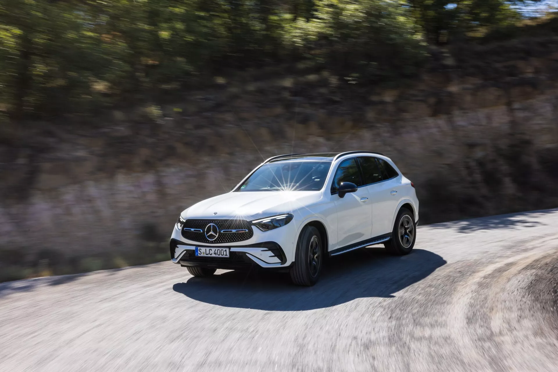 Prise dynamique d'un SUV Mercedes-Benz GLC 2022 blanc en mouvement, capturée en plein jour sous un angle de trois quarts avant. Points forts: la calandre avant avec un emblème proéminent, les phares LED distinctifs, le pare-chocs avant avec des prises d'air visibles, le capot sculpté avec des lignes surélevées et le profil latéral jusqu'aux portes arrière. Les roues sont représentées en mouvement avec un léger flou, suggérant la vitesse. Les rétroviseurs latéraux sont conçus de manière aérodynamique. L'environnement apparaît flou en raison du mouvement du véhicule, soulignant la nature dynamique et rapide de la voiture.
