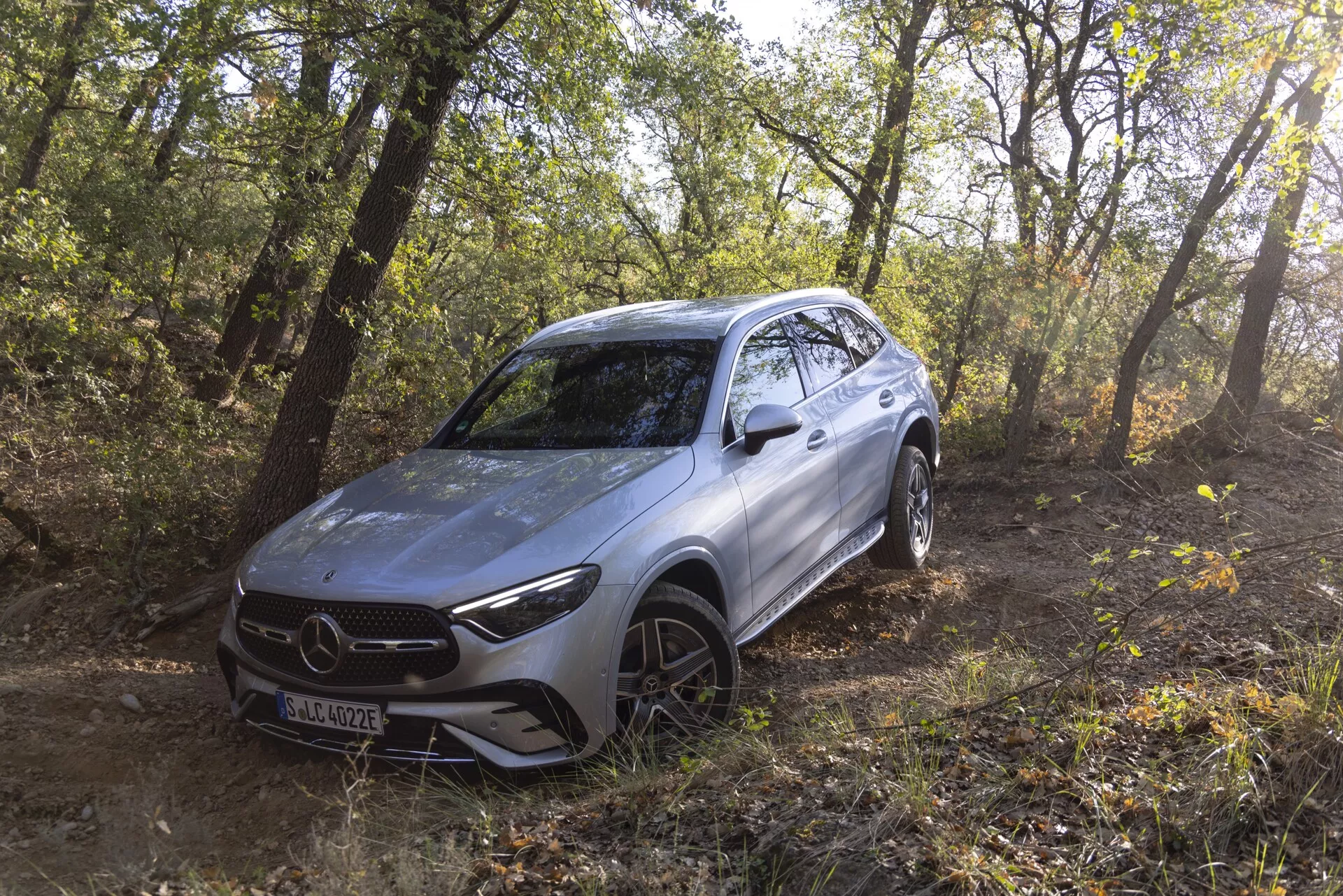 Image d'un SUV Mercedes GLC 2022 argenté positionné en plein air dans une zone boisée, montrant ses capacités hors route. Le véhicule est incliné vers la gauche, offrant une vue de trois quarts du profil avant et latéral, incluant la calandre avant, les phares LED aiguisés, le pare-chocs avant profilé, le capot, les roues en alliage avec des pneus tout-terrain et des lignes fluides. La lumière naturelle du soleil filtre à travers les arbres, soulignant le design moderne et aérodynamique et suggérant des fonctionnalités avancées d'assistance à la conduite logées derrière l'emblème de la calandre.
