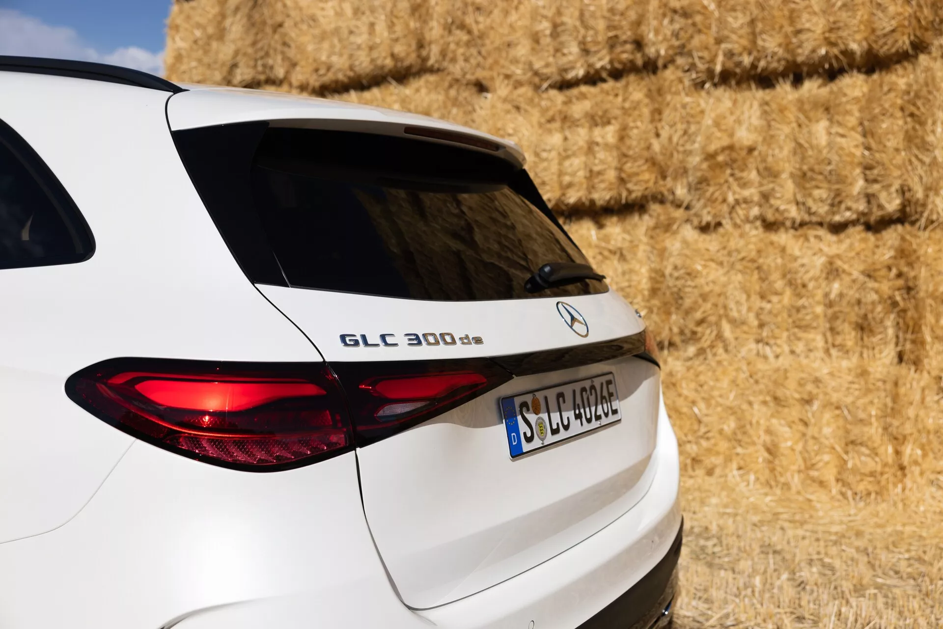 Vue arrière en trois quarts rapprochée d'un SUV Mercedes-Benz GLC 300 d blanc, montrant le côté droit de l'arrière incluant le hayon, le feu arrière allumé et une portion des fenêtres arrière. Le véhicule est stationné dans un cadre rural avec des bottes de foin en arrière-plan. Les caractéristiques visibles incluent le nom du modèle 'GLC 300 d', le logo Mercedes-Benz et une plaque d'immatriculation de format européen, mettant en avant les lignes élégantes du design et l'éclairage moderne du véhicule.