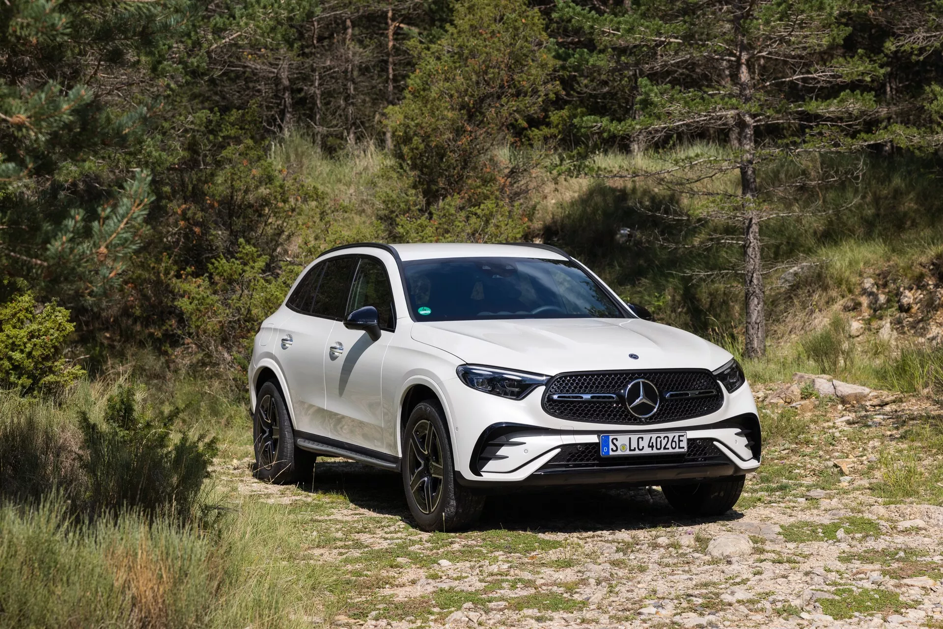 Image du SUV Mercedes-Benz GLC 2022 dans un cadre naturel, positionné sur un chemin de terre ou de gravier avec de la végétation en arrière-plan. Le véhicule est capturé en vue de trois quarts avant, montrant la calandre avant avec un grand emblème, des phares à LED nets et une partie de la carrosserie latérale. Le SUV dispose de jantes en alliage noires à plusieurs rayons, de rétroviseurs latéraux avec clignotants intégrés et d'une ligne de ceinture ascendante menant à une arrière sportif et effilé. La carrosserie est de couleur blanche unie, et l'image suggère que ce modèle fait partie de la gamme électrifiée de Mercedes-Benz, comme l'indique le badge 'EQ' sur l'aile avant.