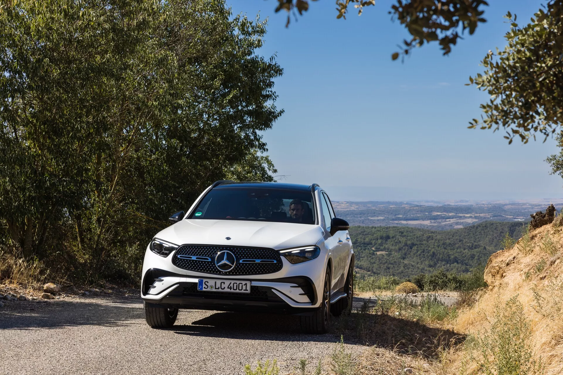 Une Mercedes-Benz GLC 2022 moderne est positionnée sur une route étroite avec un arrière-plan naturel et vallonné surplombant une vallée ou des plaines. L'emblème avant et le design de la grille de la voiture sont clairement visibles, capturés d'une perspective frontale basse légèrement décalée sur le côté gauche, mettant en valeur la grille avant, les phares et une partie du capot. Les phares à LED, une grille distinctive avec un grand emblème et un design de pare-chocs sportif mettent en évidence les caractéristiques avancées et élégantes de la voiture. La prise de vue de jour avec un éclairage naturel évoque la tranquillité, montrant les systèmes avancés d'assistance à la conduite suggérés par l'emblème. Le profil aérodynamique élégant est accentué par les lignes du capot qui se fondent avec les côtés sculptés, capturé à une distance d'environ 3-5 mètres de la caméra.