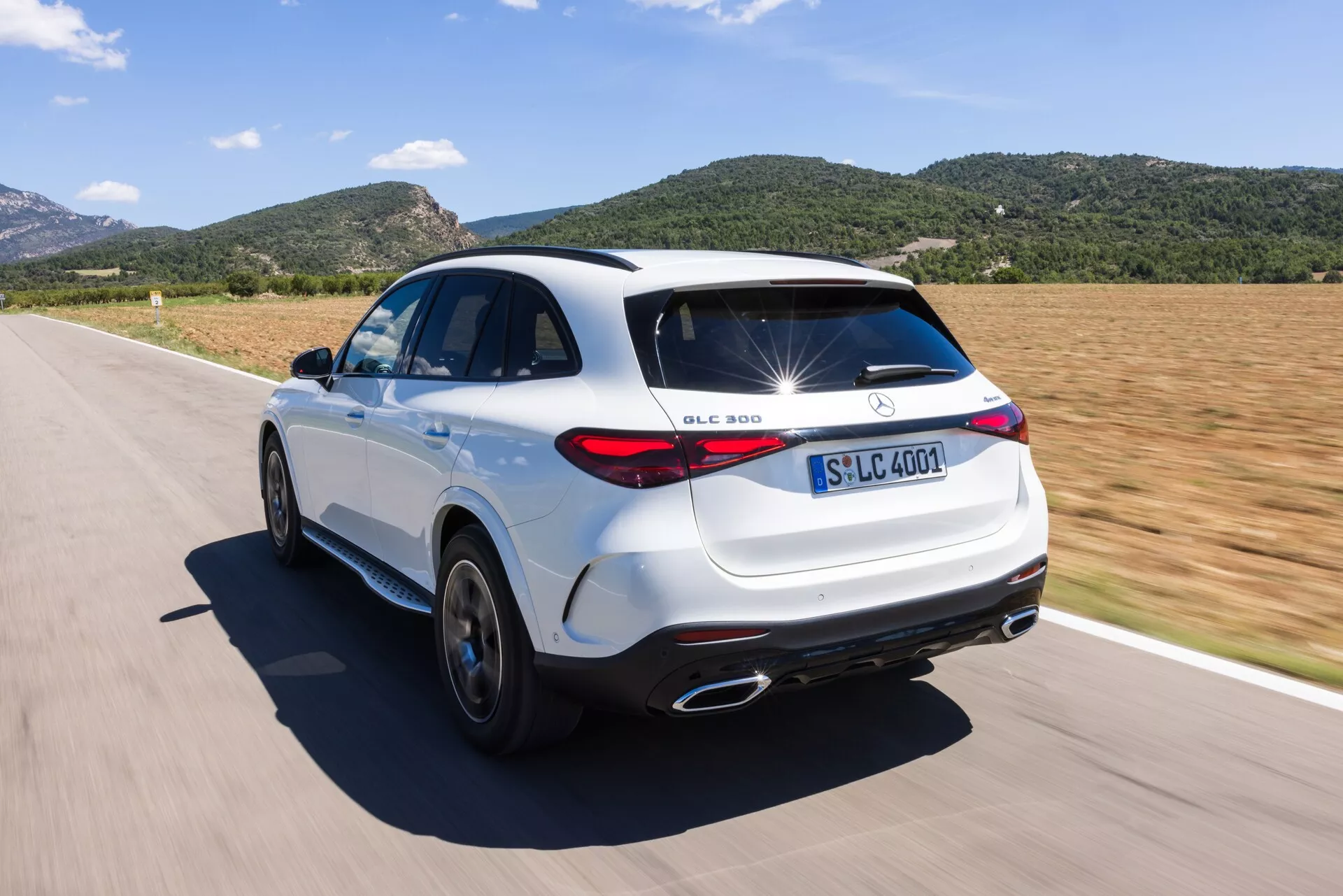 Vue en angle trois quarts arrière haut d'un SUV Mercedes-Benz GLC 300 blanc sur une route. L'image met en avant des éléments de design comme les badges identifiables, les feux arrière à LED, les embouts d'échappement chromés, les jantes en alliage à cinq branches et le becquet de toit distinctif. Le SUV est en mouvement par une journée ensoleillée, soulignant ses capacités dynamiques et son esthétique sportive.