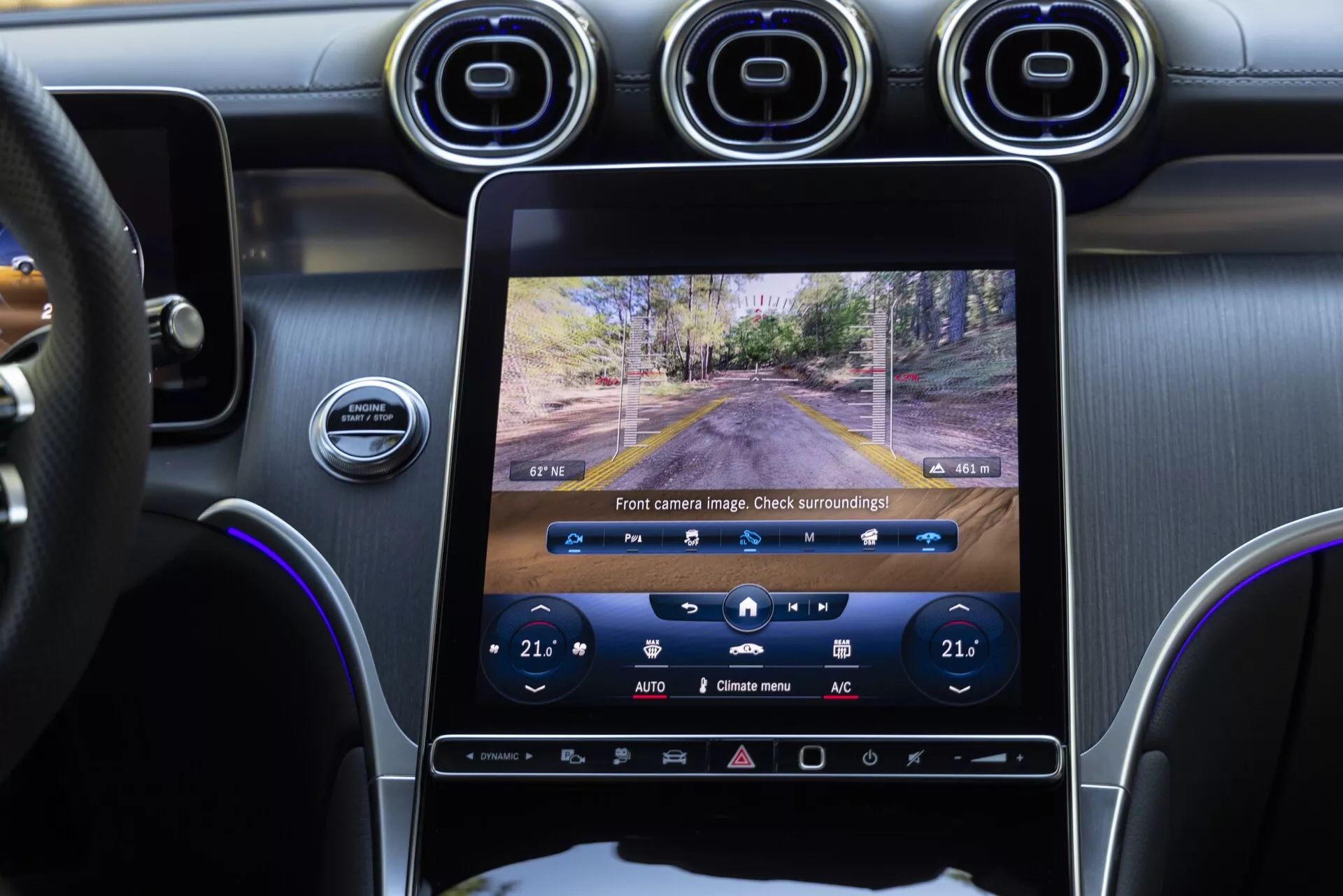 Vue intérieure de la Mercedes GLC 2022 centrée sur la console centrale et le système d'infodivertissement, prise depuis la perspective du passager arrière. Le grand écran tactile vertical affiche une vue en direct de la caméra frontale, avec les commandes de climatisation réglées à 21°C visibles en dessous. Le tableau de bord, le volant et une partie de la porte du conducteur sont également visibles, montrant une combinaison élégante d'interfaces numériques et de boutons physiques, des bouches d'aération circulaires métalliques et un volant en cuir.