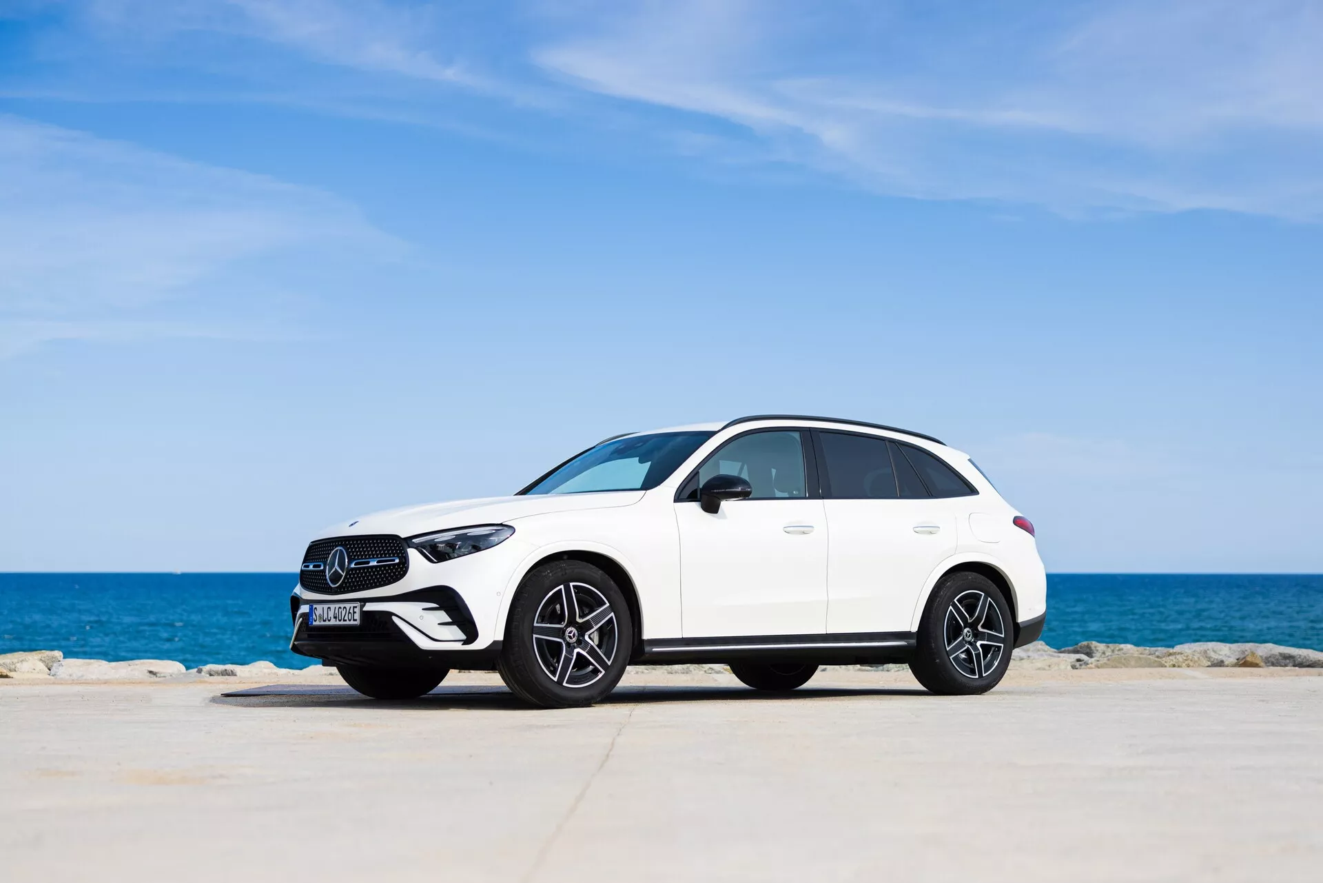 Vue de profil latéral d'un SUV Mercedes GLC 2022 blanc moderne, garé devant un arrière-plan pittoresque avec un ciel bleu clair et un horizon marin calme. Le véhicule, bien centré dans le cadre, présente des courbes élégantes et aérodynamiques, des roues en alliage multi-branches et des phares à LED. La calandre avant et le design de la roue avant sont subtilement mis en valeur, offrant un mélange de luxe et de sportivité dans un environnement paisible.