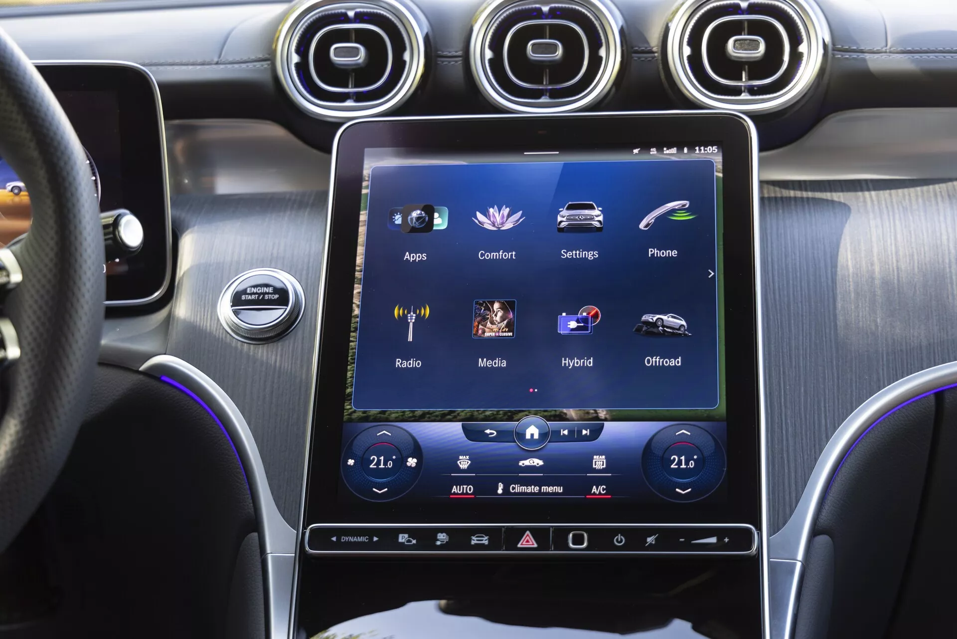 La console centrale d'une Mercedes-Benz GLC 2022 vue depuis le siège du conducteur, focalisée sur le grand écran tactile vertical du système d'infodivertissement affichant des options comme 'Apps,' 'Confort,' 'Réglages,' avec des commandes de climatisation bi-zone, des aérations métalliques, un bouton de démarrage et un mélange premium de finitions métalliques brossées et noir brillant.