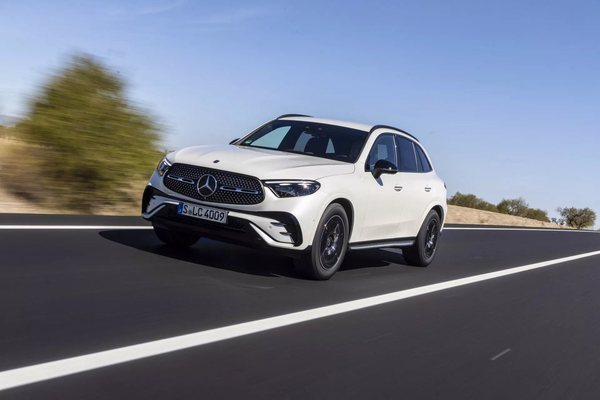 Mercedes GLC 2022 (254) crossover SUV blanc à quatre portes en mouvement sur une route ouverte, capturé sous un angle bas mettant en avant sa hauteur et sa posture. Le véhicule se déplace en diagonale de gauche à droite, avec un accent sur la calandre avant, les phares, les feux de jour à LED et le pare-chocs avant inférieur. Le profil latéral révèle la ligne sculptée du toit, les lignes de design, les arches de roue évasées et les jantes en alliage, combinant luxe et sportivité. Le flou d'arrière-plan suggère la vitesse, sous un ciel dégagé de jour.
