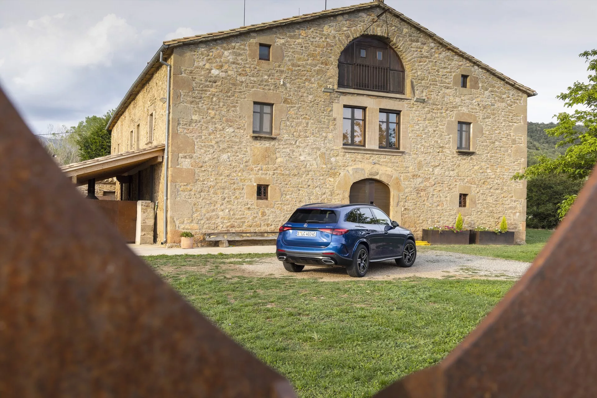 Mercedes GLC 2022 (254) garé sur une zone herbeuse avec une maison en pierre traditionnelle en arrière-plan, montrant le profil arrière et partiel latéral avec des feux arrière à LED, un becquet de toit, des rails de toit, des capteurs de stationnement arrière, des jantes en alliage et des éléments en fer rouillé au premier plan.