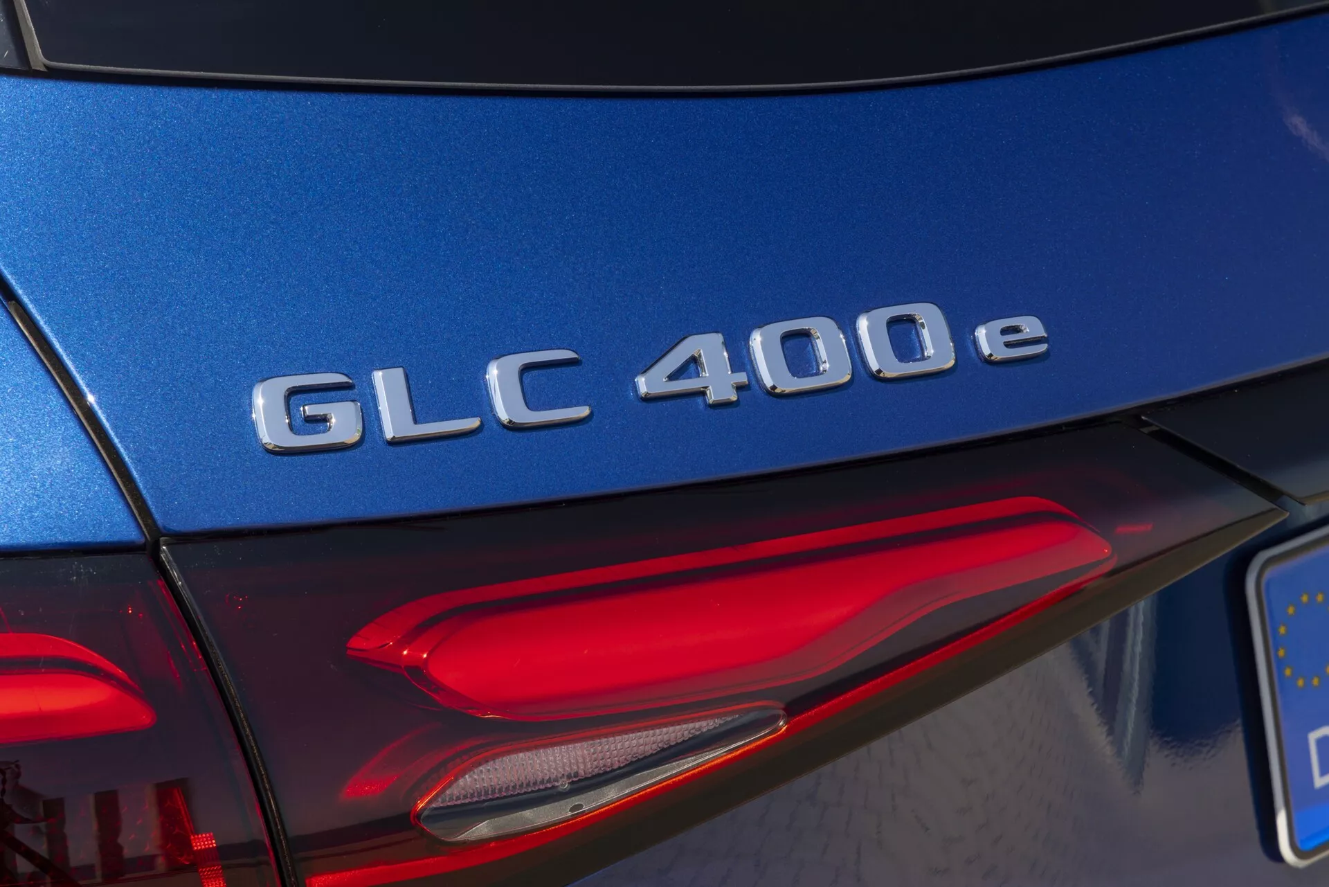 Détail de la section arrière d'une Mercedes-Benz GLC 400e 2022, avec un gros plan sur la désignation du modèle en chrome sur le hayon bleu, avec une prise de vue en angle diagonal capturant une partie de l'assemblage du feu arrière gauche avec des éléments LED en plein jour.
