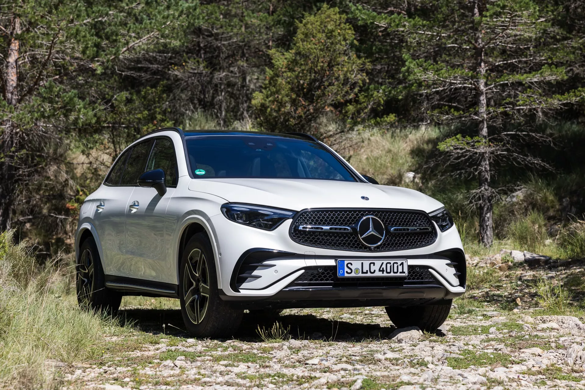 Vue de trois-quarts avant d'une Mercedes-Benz GLC 2022, mettant en évidence sa calandre proéminente, ses phares LED élégants, ses grandes roues en alliage noir à plusieurs rayons et son corps aérodynamique blanc. Le véhicule est garé sur un terrain rocheux avec des arbres en arrière-plan, soulignant ses capacités de SUV et son attrait luxueux.