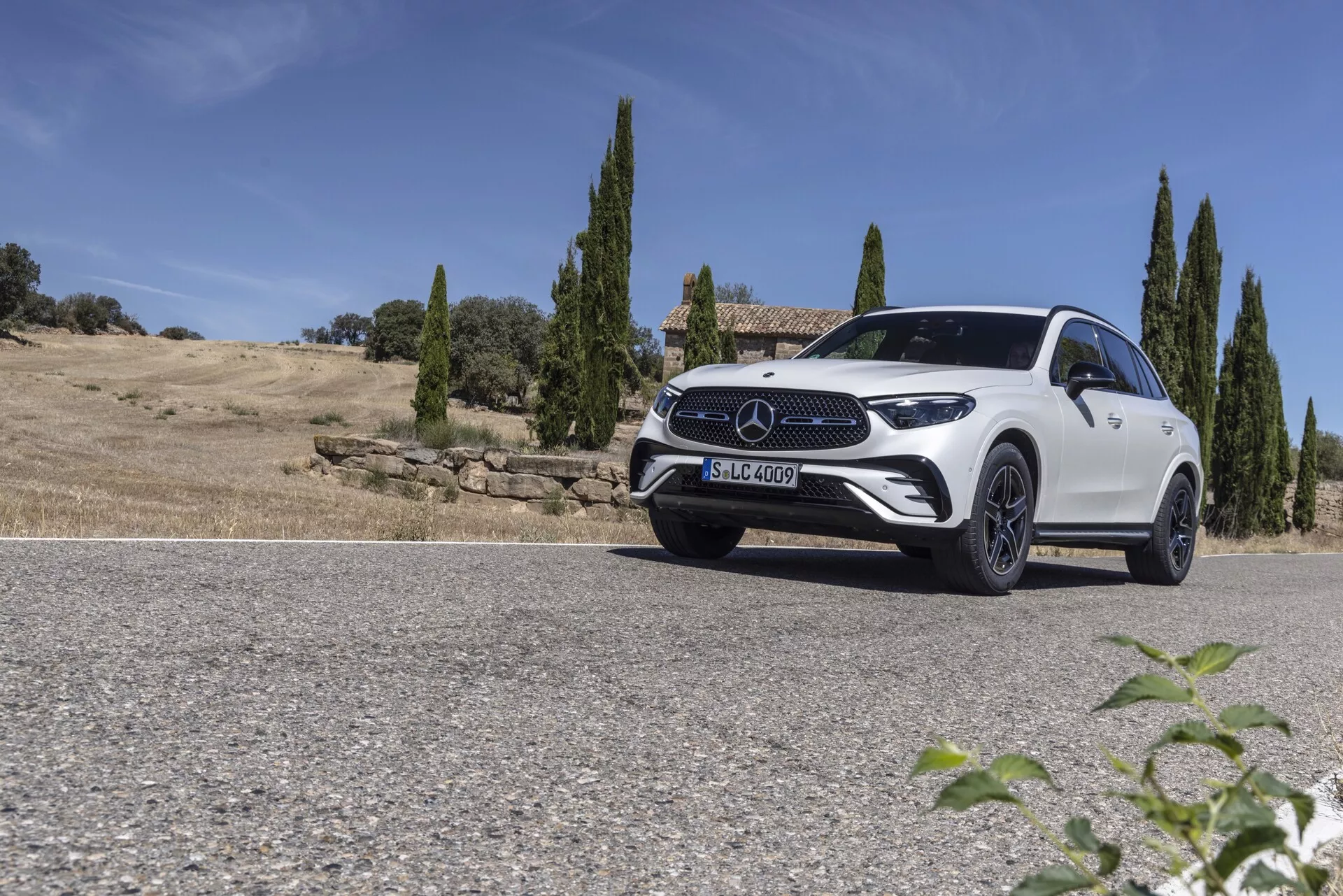 Prise de vue en contre-plongée d'un SUV Mercedes GLC 2022 blanc garé sur une route contre un paysage naturel avec un ciel clair et des cyprès en arrière-plan. Le quart avant du véhicule est capturé du côté du conducteur, montrant les feux LED élégants, la calandre avant proéminente avec un grand emblème, les roues en alliage sport et le capot profilé. La ligne de toit légèrement inclinée et la ligne d'épaule bien définie mettent en valeur le design du SUV combinant aérodynamisme et robustesse. La lumière du soleil brillante crée des ombres et des reflets, rehaussant l'esthétique luxueuse et moderne de la voiture.