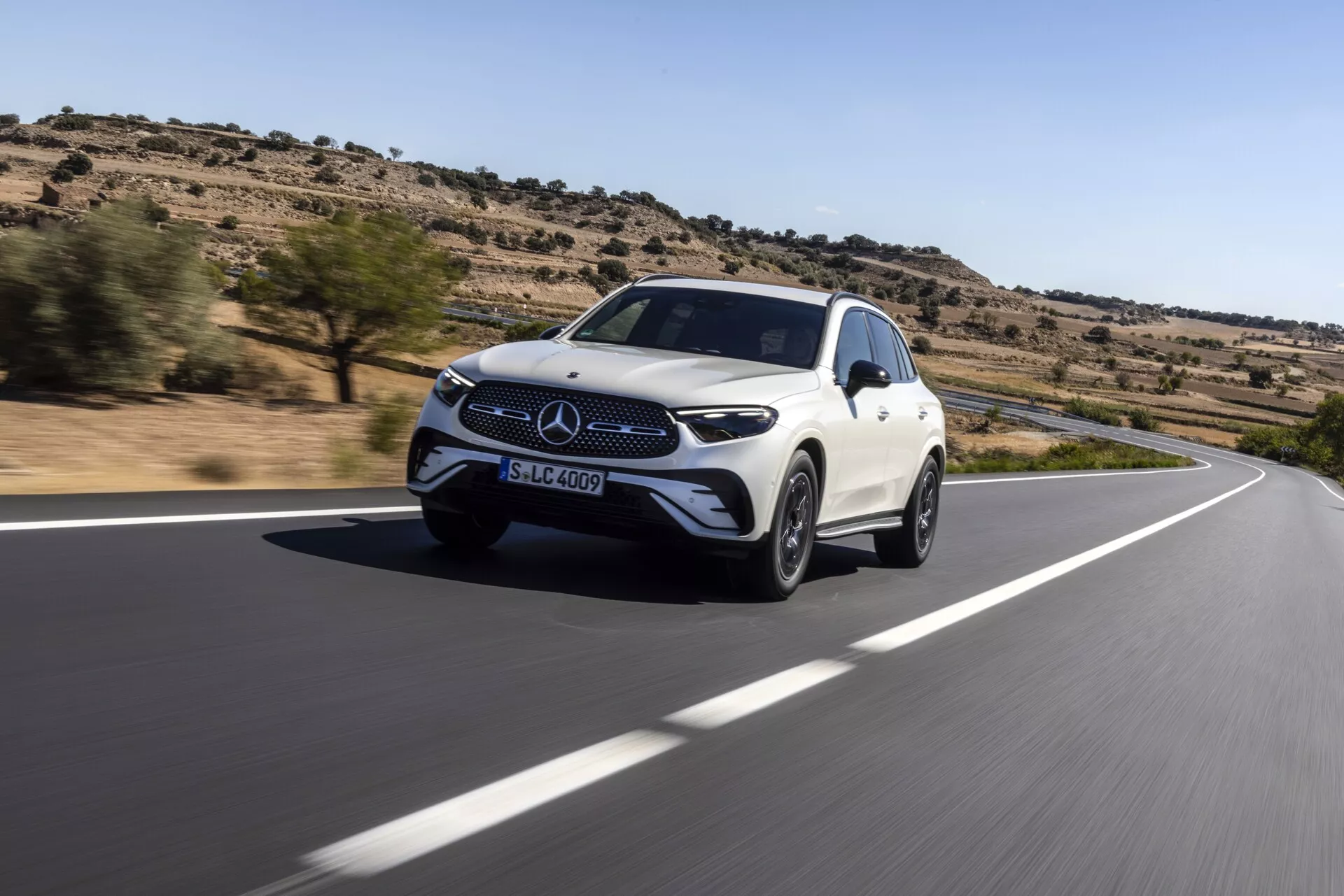 Photo dynamique à un angle bas d'un SUV Mercedes-Benz GLC blanc 2022 en mouvement sur une route sinueuse. La vue de trois quarts avant montre la calandre avant avec l'emblème Mercedes-Benz, les phares et les ailes avant. Le design aérodynamique et les jantes en alliage soulignent la vitesse et le luxe du véhicule. Vu dans un environnement désertique avec une végétation clairsemée, les capteurs avant intégrés et les rétroviseurs latéraux de la voiture suggèrent des systèmes avancés d'assistance à la conduite et de sécurité.
