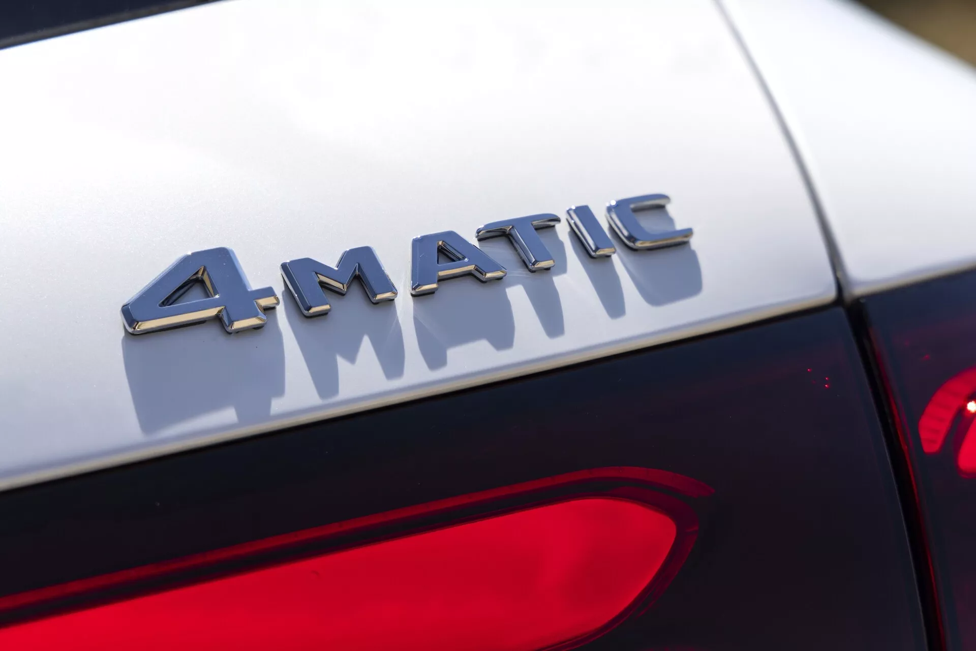 Vue rapprochée du badge 4MATIC à l'arrière d'une Mercedes GLC 2022. La photographie capture le badge en mise au point nette avec des lettres métalliques réfléchissant la lumière, montées juste au-dessus du feu arrière. L'arrière-plan est flou, mettant en valeur l'effet tridimensionnel du badge et le contraste avec la peinture sombre de la voiture. Conception du feu arrière partiellement visible dans le coin inférieur droit.