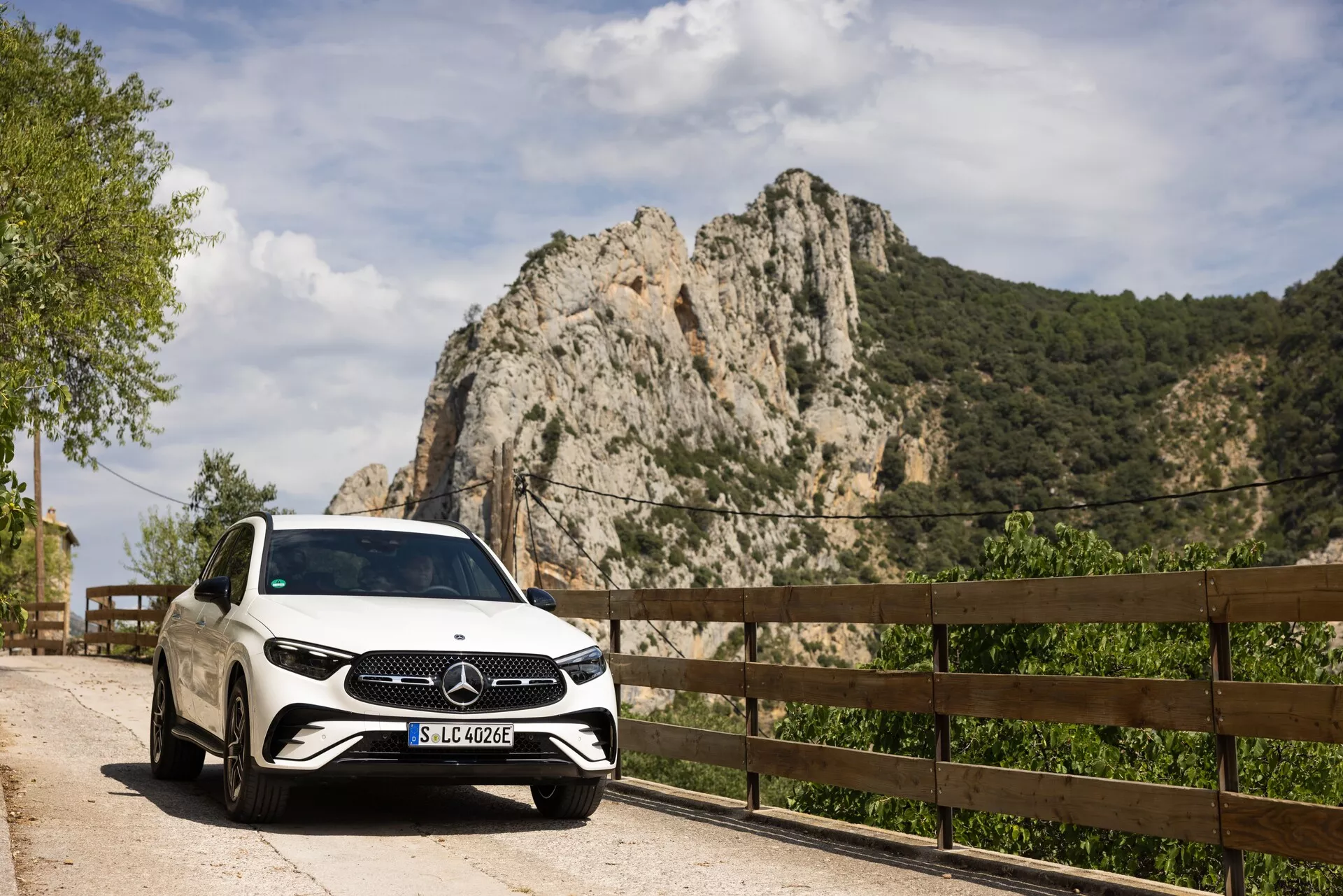 Voiture blanche Mercedes-Benz GLC 2022 garée sur un bord de route pittoresque avec un angle de vue avant-droit montrant la calandre, les phares et une partie du côté droit, y compris la porte passager avant et la roue. Plaque d'immatriculation 'SLC 400 e' indiquant un possible véhicule hybride rechargeable ou électrique. Photographié en plein jour, la peinture blanche et la surface brillante de la voiture reflètent la lumière naturelle, mettant en valeur le grand emblème Mercedes-Benz sur la calandre. Montagne rocheuse et garde-corps en bois en arrière-plan suggèrent une route rurale ou de montagne, évoquant un retrait luxueux ou une exploration en plein air.