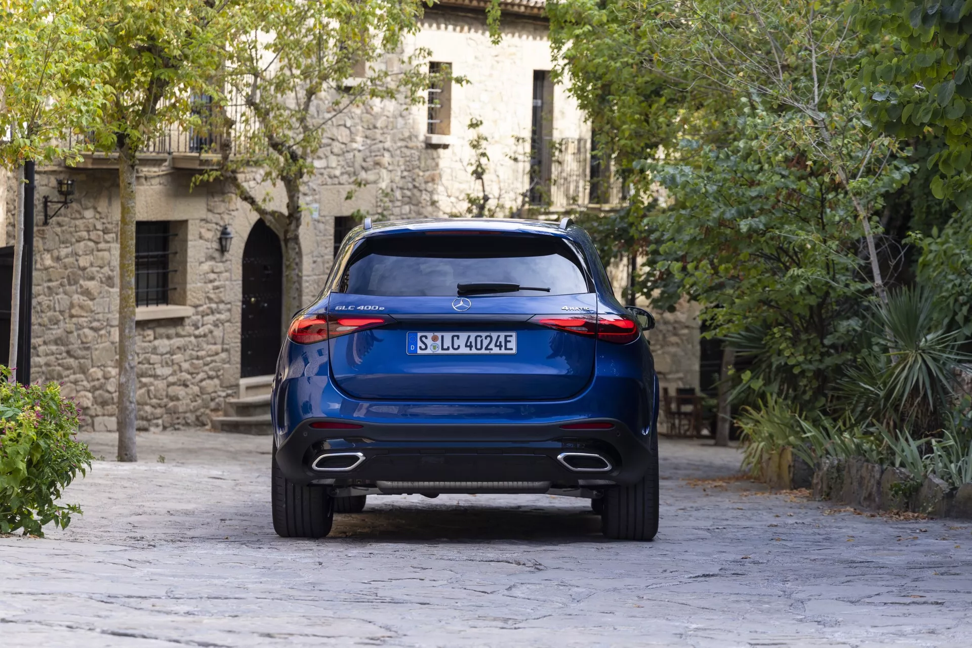 Vue de l'angle arrière d'un SUV Mercedes GLC 2022 bleu sous une perspective légèrement basse, montrant sa hauteur de caisse élevée, ses sorties d'échappement doubles et ses feux arrière élégants. Le véhicule, positionné au centre, se dresse sur un arrière-plan d'architecture européenne et de pavé en pierre, mettant en avant sa modernité et son possible groupe motopropulseur électrique.