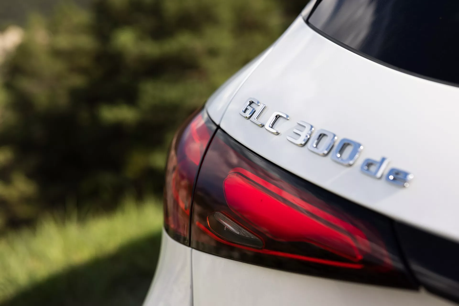 Vue rapprochée de la section arrière de la Mercedes GLC 300de 2022, avec un focus sur le badge du modèle et une partie du feu arrière à LED. L'image, prise sous un angle latéral légèrement surélevé, met en évidence le badge GLC 300de et le contour lisse du design moderne du feu arrière. La peinture métallique reflète subtilement la lumière, suggérant une surface bien entretenue, bien que la couleur exacte semble être claire, probablement blanche ou argentée. La faible profondeur de champ maintient le badge en nette focale tout en floutant l'arrière-plan et les zones périphériques, mettant en valeur les détails technologiques et de design de ce véhicule hybride diesel-électrique.