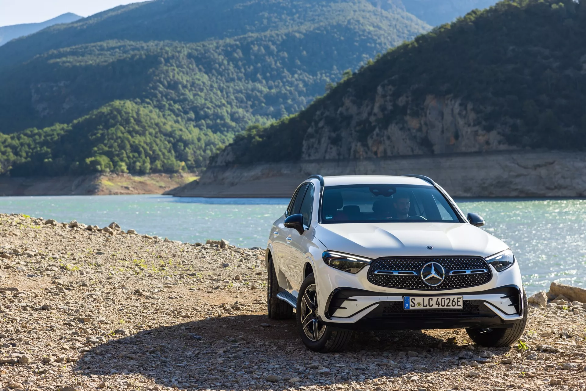 Vue de trois quarts avant d'une voiture de luxe Mercedes GLC 254 de 2022, mettant en avant sa calandre avant avec un emblème proéminent, des phares LED élégants et des jantes en alliage, garée dans un cadre extérieur accidenté près du rivage avec des montagnes en arrière-plan.