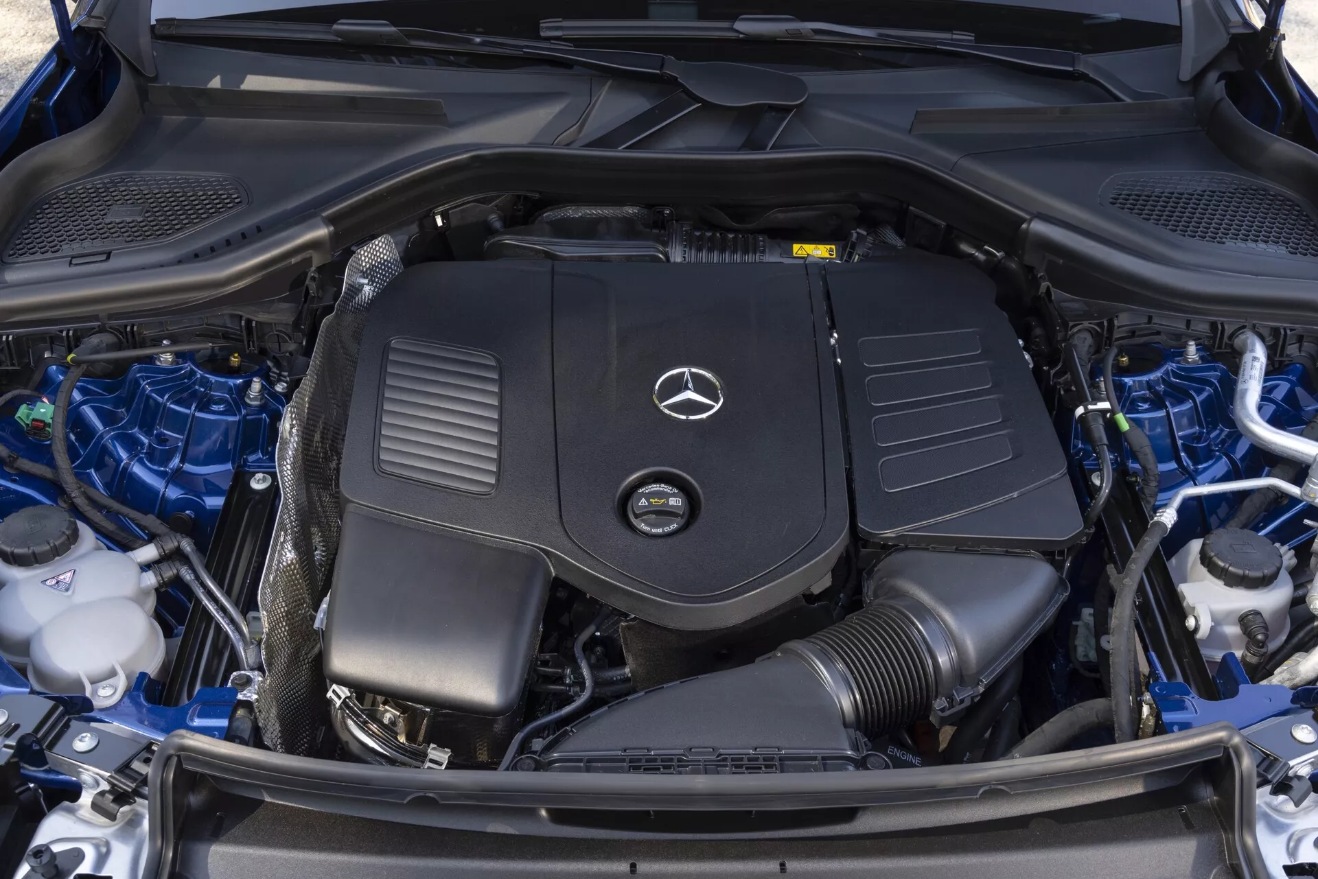 Vue en gros plan de haut en bas du compartiment moteur propre et bien entretenu d'une Mercedes GLC 2022, montrant le cache moteur avec le logo Mercedes-Benz bien en évidence, la batterie, les réservoirs de liquide, les composants du système de refroidissement et les faisceaux de câbles soigneusement organisés.
