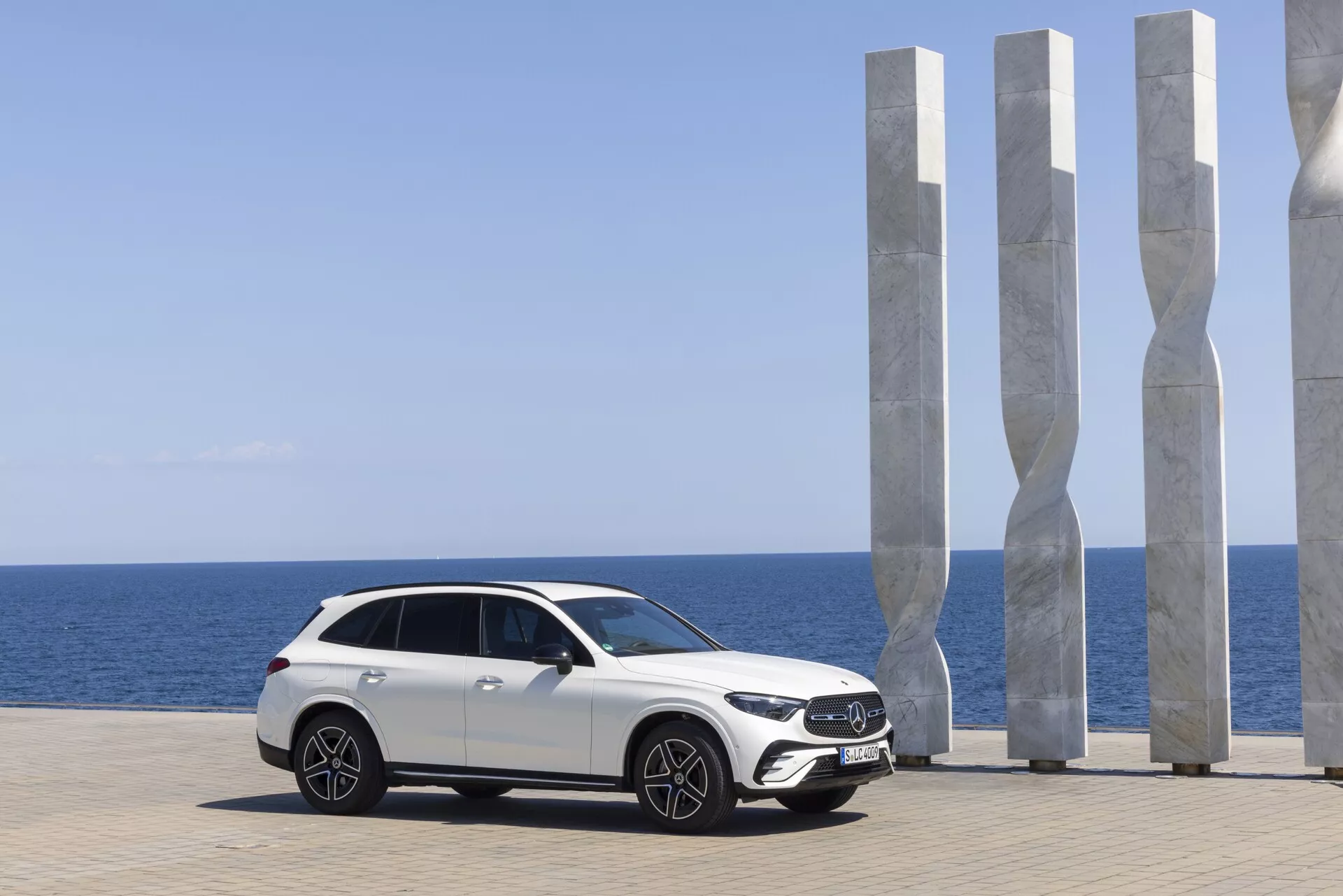 Mercedes GLC 2022 blanc moderne et luxueux garé avec vue côtière pittoresque; profil latéral mettant en valeur un design élégant avec garde au sol surélevée, grandes roues en alliage, phares LED avancés, rétroviseurs aérodynamiques avec clignotants intégrés, ligne de toit inclinée façon coupé, toit panoramique et vitres teintées. Arrière-plan pittoresque avec sculptures blanches modernes, ciel bleu clair et mer calme rehaussant l'attrait automobile haut de gamme.