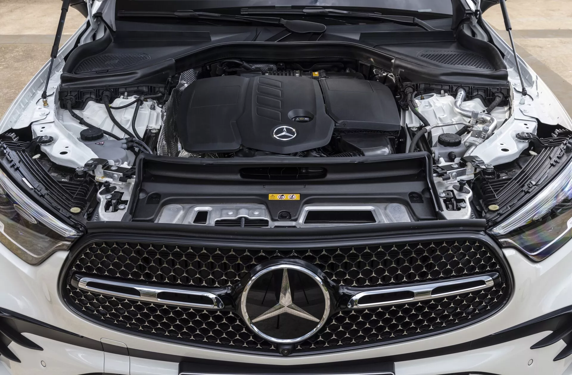 Vue de face rapprochée de la Mercedes-Benz GLC 2022 (254) avec le capot ouvert montrant le compartiment moteur depuis une perspective au niveau des yeux centrée sur l'emblème de la marque et la calandre, soulignant la grande calandre à motif diamanté, l'emblème étoilé de Mercedes-Benz, le compartiment moteur avec le logo et des composants visibles comme le bloc moteur, le système d'admission d'air, le réservoir de liquide de refroidissement, la tour de suspension et la caméra frontale, indiquant des caractéristiques technologiques avancées.