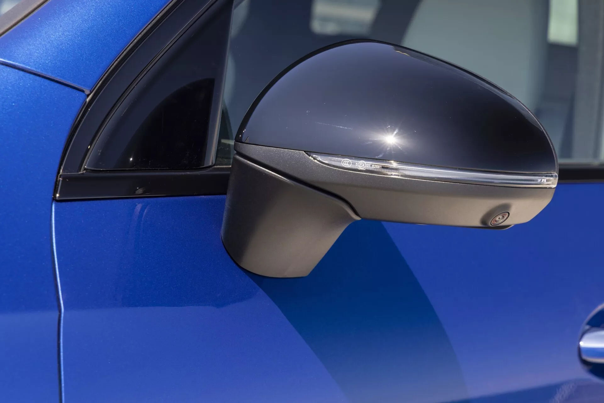 Gros plan du rétroviseur latéral d'une Mercedes GLC 2022 (254) bleu vif, montrant le côté conducteur avec un design argenté/chromé profilé et un clignotant à LED intégré. La photo, prise en perspective latérale avec le rétroviseur net et l'arrière-plan flou, souligne les caractéristiques aérodynamiques et luxueuses du design moderne de la voiture.