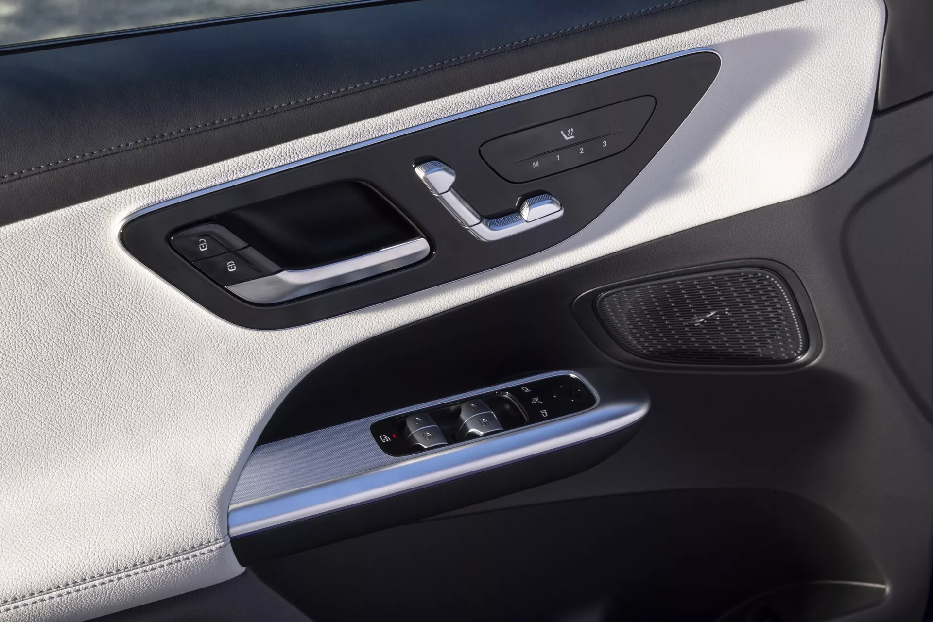 Vue en gros plan du panneau intérieur de la porte de la Mercedes GLC 2022 254, centrée sur le côté conducteur ou passager, avec poignée argentée et contrôle de verrouillage intégré, boutons de mémoire du siège, interrupteurs de fenêtre électrique, joystick pour les réglages du rétroviseur latéral, grille de haut-parleur, matériaux luxueux comprenant une section supérieure en similicuir, accents argentés et éclairage ambiant.