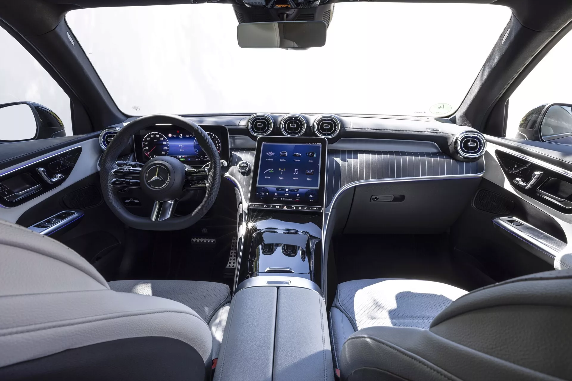 Intérieur du Mercedes GLC 2022, vu depuis le siège central arrière. L'habitacle luxueux présente une palette de couleurs bicolore avec du cuir crème et des accents sombres. Le tableau de bord est doté d'un élégant décor en bois avec un grand système d'infodivertissement à écran tactile vertical, des aérateurs et des boutons de commande. On peut également voir le volant multifonction en cuir avec des accents métalliques et le tableau de bord numérique.