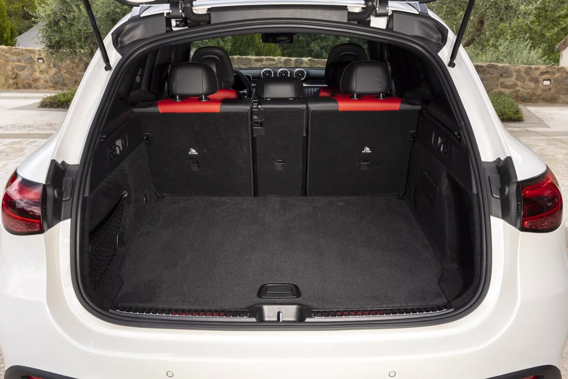 Vue arrière de l'espace de coffre du Mercedes GLC 2022 avec le hayon ouvert, présentant un sol plat revêtu de moquette, des garnitures en plastique sur les côtés et les sièges arrière rabattables avec répartition 60/40 pour agrandir l'espace de chargement. Points forts : boucles de ceinture de sécurité rouges exposées, mécanismes de pliage des sièges, points d'ancrage, et une zone encastrée pour un stockage supplémentaire.