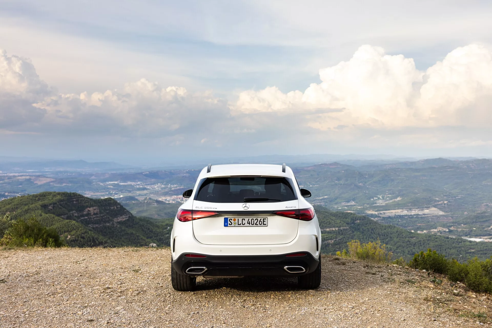 Vue arrière d'un SUV hybride/électrique Mercedes-Benz GLC 2022 (EQ) blanc, stationné sur un sol de gravier avec un panorama de montagnes et d'arbres sous un ciel partiellement nuageux. La photo met en avant les feux arrière à LED horizontaux, les doubles sorties d'échappement, le diffuseur arrière et le marquage EQ. La composition suggère des thèmes d'aventure et de voyage écologique.