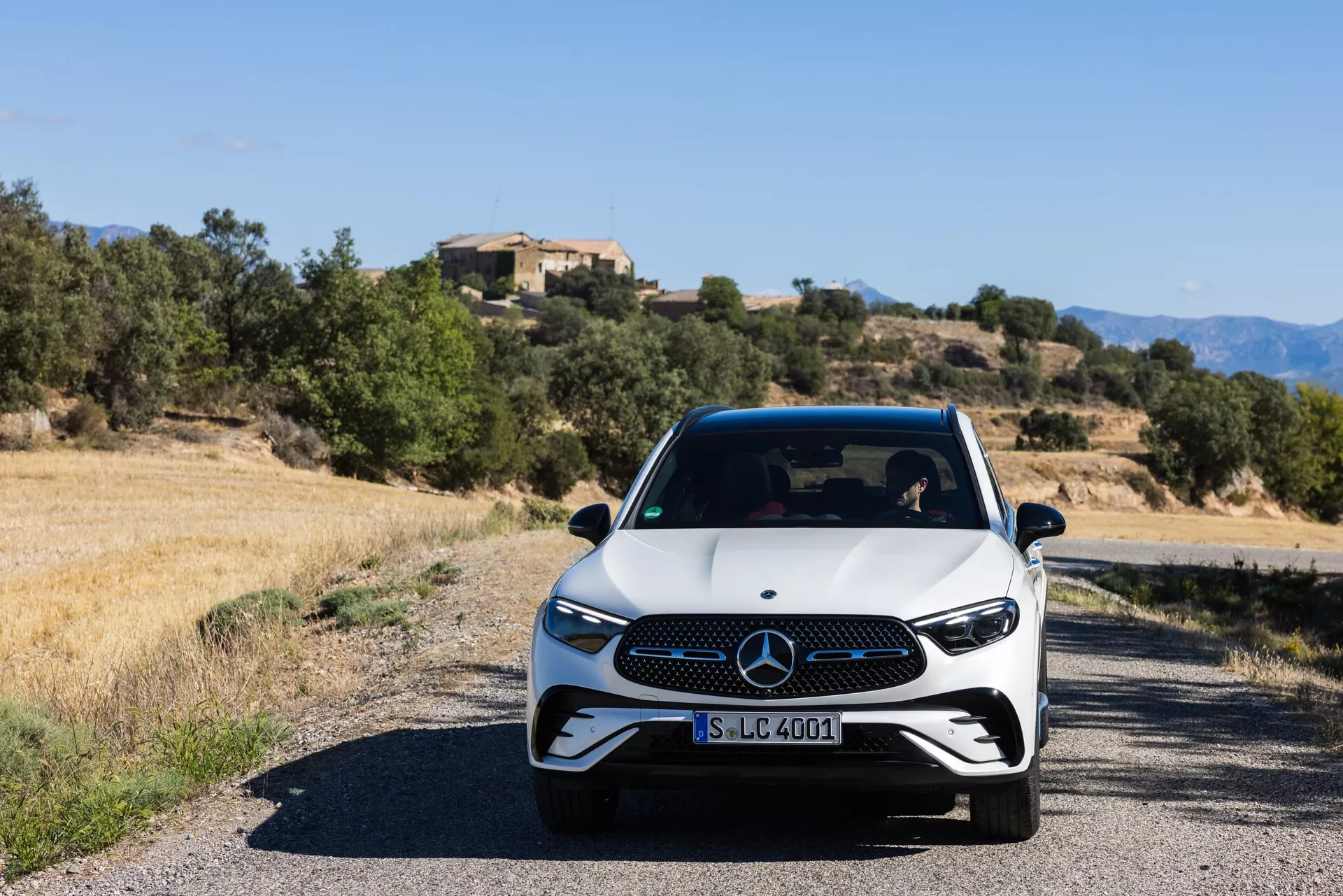 Vue de face d'une Mercedes-Benz GLC 2022 (254) moderne sur une route étroite, avec une base blanche et des détails noirs, des phares LED et une grille distinctive. La photo prise à hauteur des yeux en plein jour montre la finition sportive et premium du véhicule avec un paysage méditerranéen en arrière-plan.