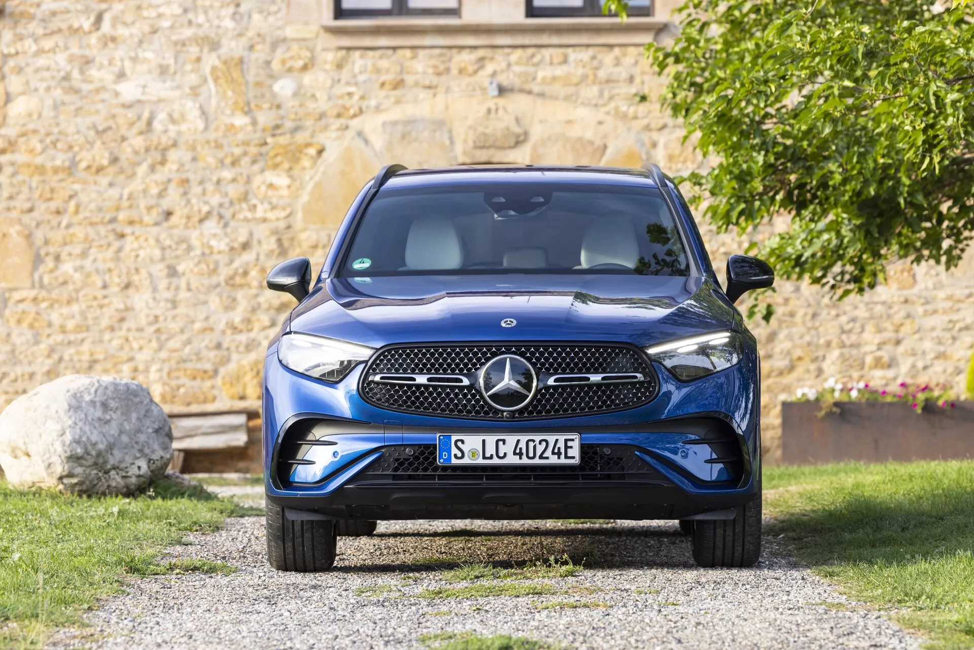 Vue frontale d'une Mercedes GLC 2022 (254) montrant la grille avant proéminente, le pare-chocs, le capot, les phares et le pare-brise. La voiture est positionnée sur une allée dans un environnement suburbain avec de la verdure et une partie d'un bâtiment en arrière-plan. La grille a une surface lisse avec un emblème qui contient probablement des capteurs ou une caméra. Les phares élégants semblent avoir une technologie LED. Le pare-chocs inférieur comprend des prises d'air et des capteurs de stationnement. L'accent bleu sur la plaque d'immatriculation indique qu'il s'agit d'un véhicule électrique (VE) avec des caractéristiques spécifiques aux VE. Le grand pare-brise offre une bonne visibilité et inclut probablement des essuie-glaces sensibles à la pluie et un capteur de lumière. Les rétroviseurs latéraux, bien que pas entièrement visibles, sont montés sur les portes et peuvent inclure la surveillance des angles morts. Les roues ont un design aérodynamique pour améliorer l'efficacité de l'autonomie. L'image est bien éclairée par la lumière naturelle, améliorant les détails et la couleur de la voiture.